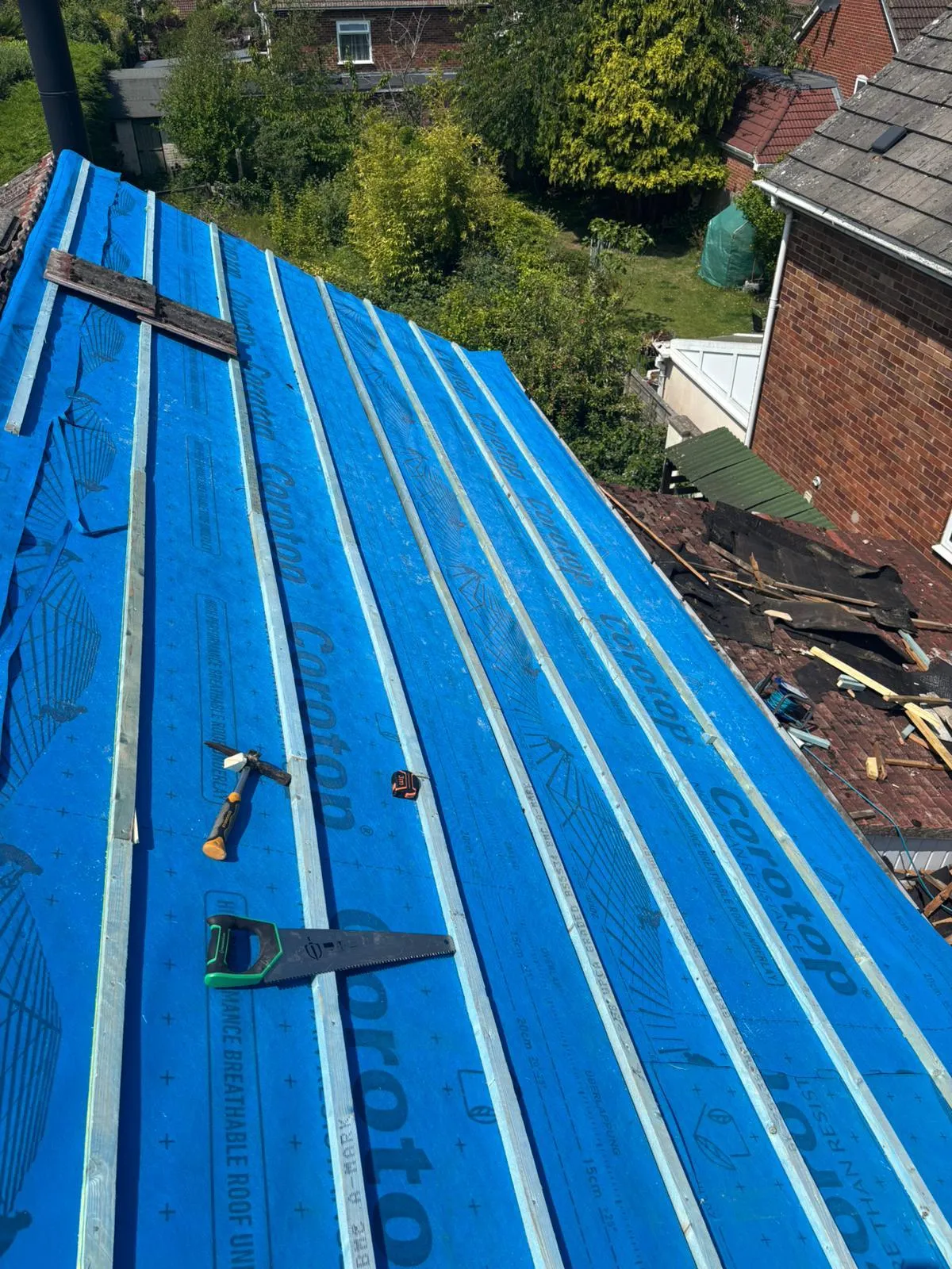 New roof installation in progress, featuring a blue breathable membrane and wooden battens on a pitched roof, with roofing tools laid out.