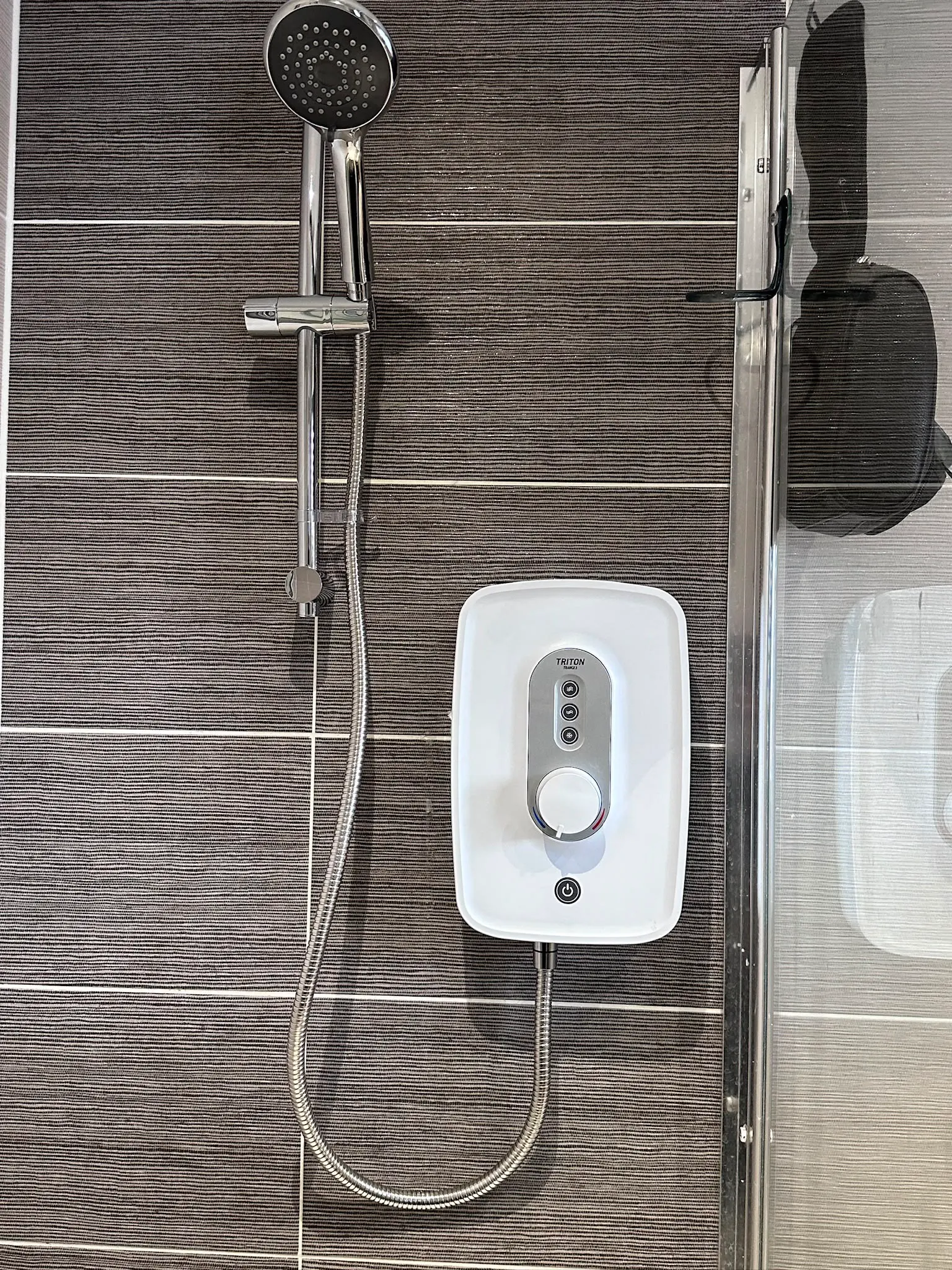 Professional electric shower installation featuring a white Triton unit and chrome fixtures on dark textured tiles.