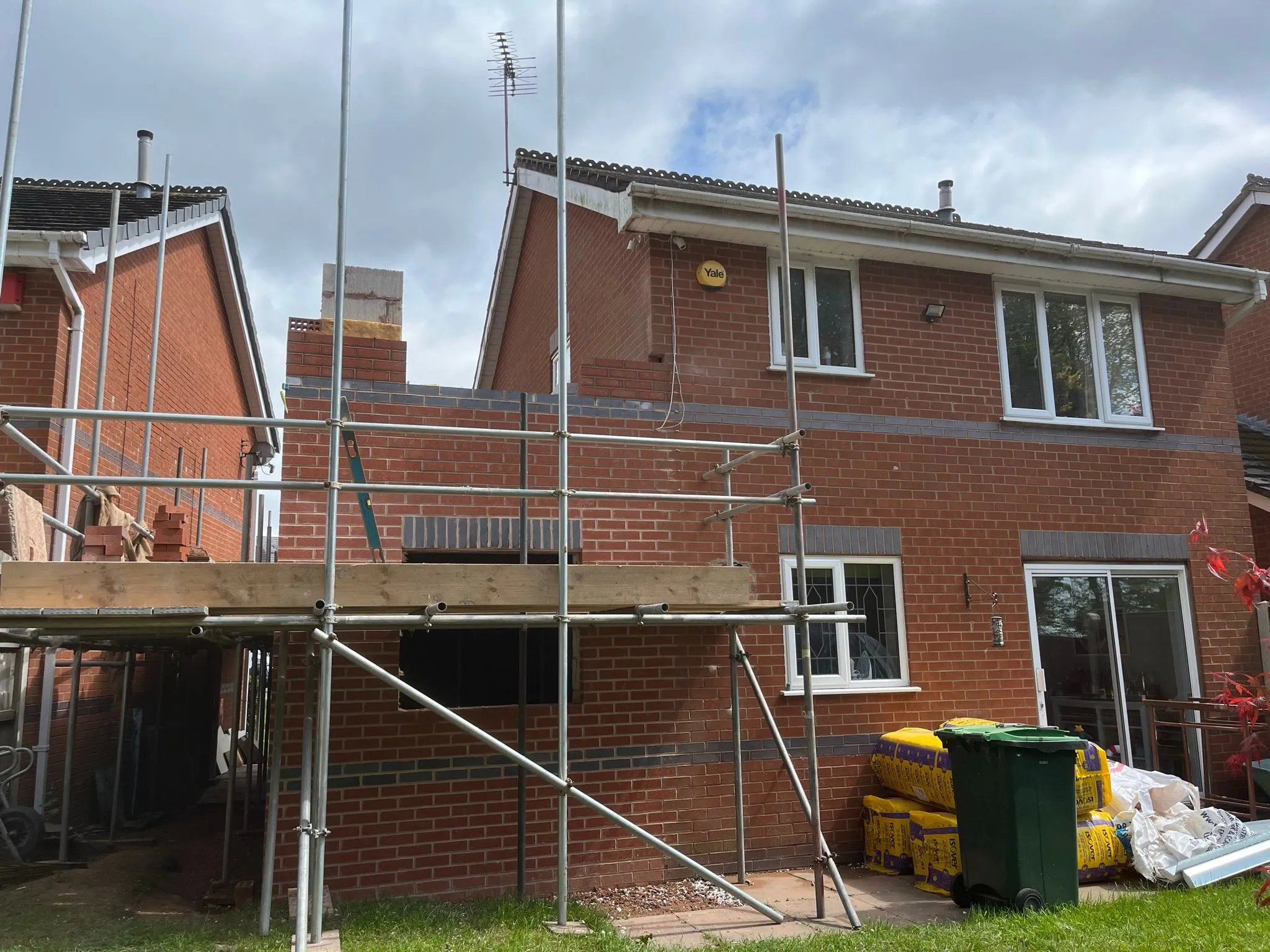 Scaffolding erected around a partially built brick home extension, with new walls, window openings, and construction materials on a cloudy day.