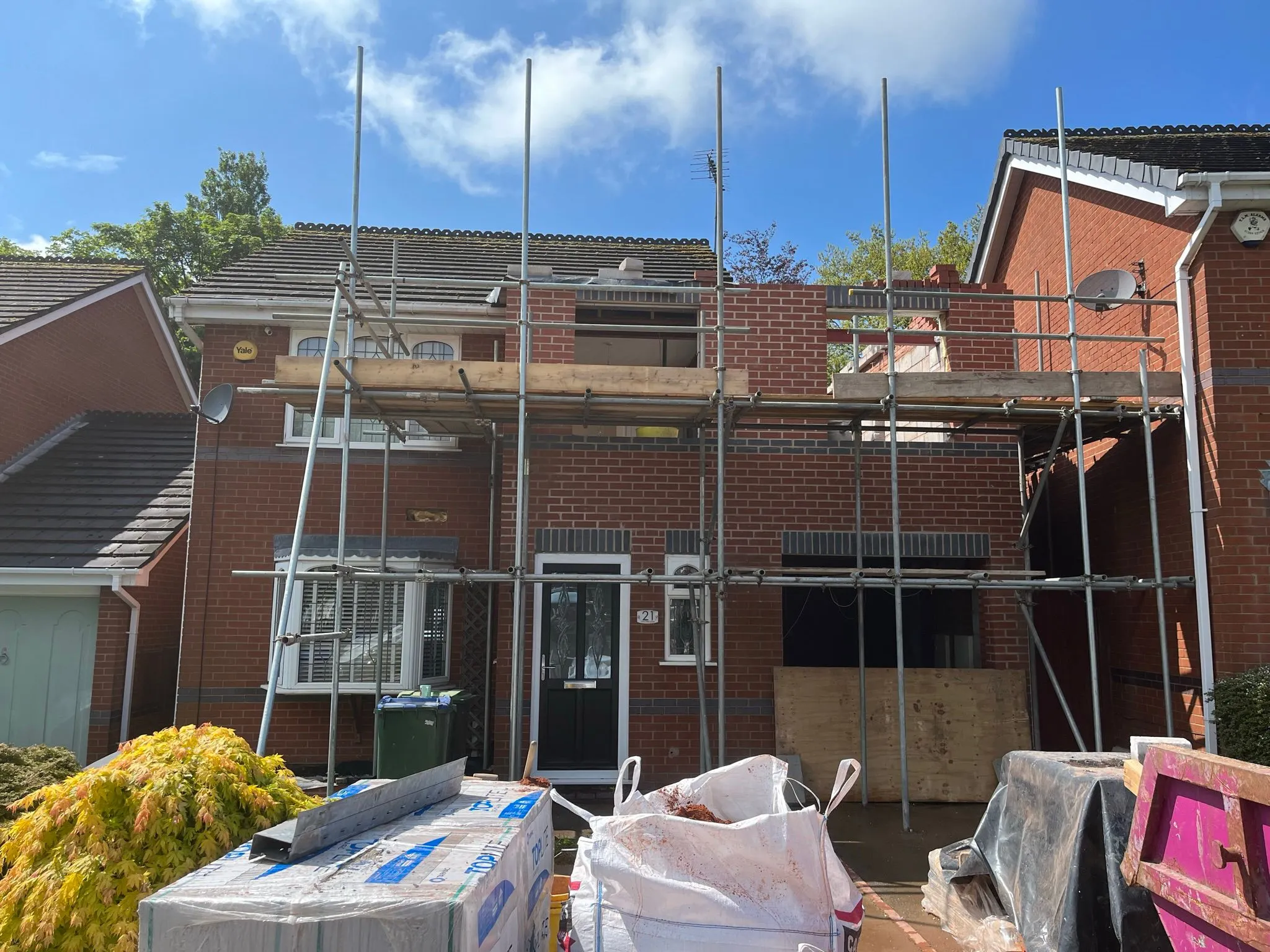 A red brick house with scaffolding and partially built walls for a new extension, with building materials in the foreground.