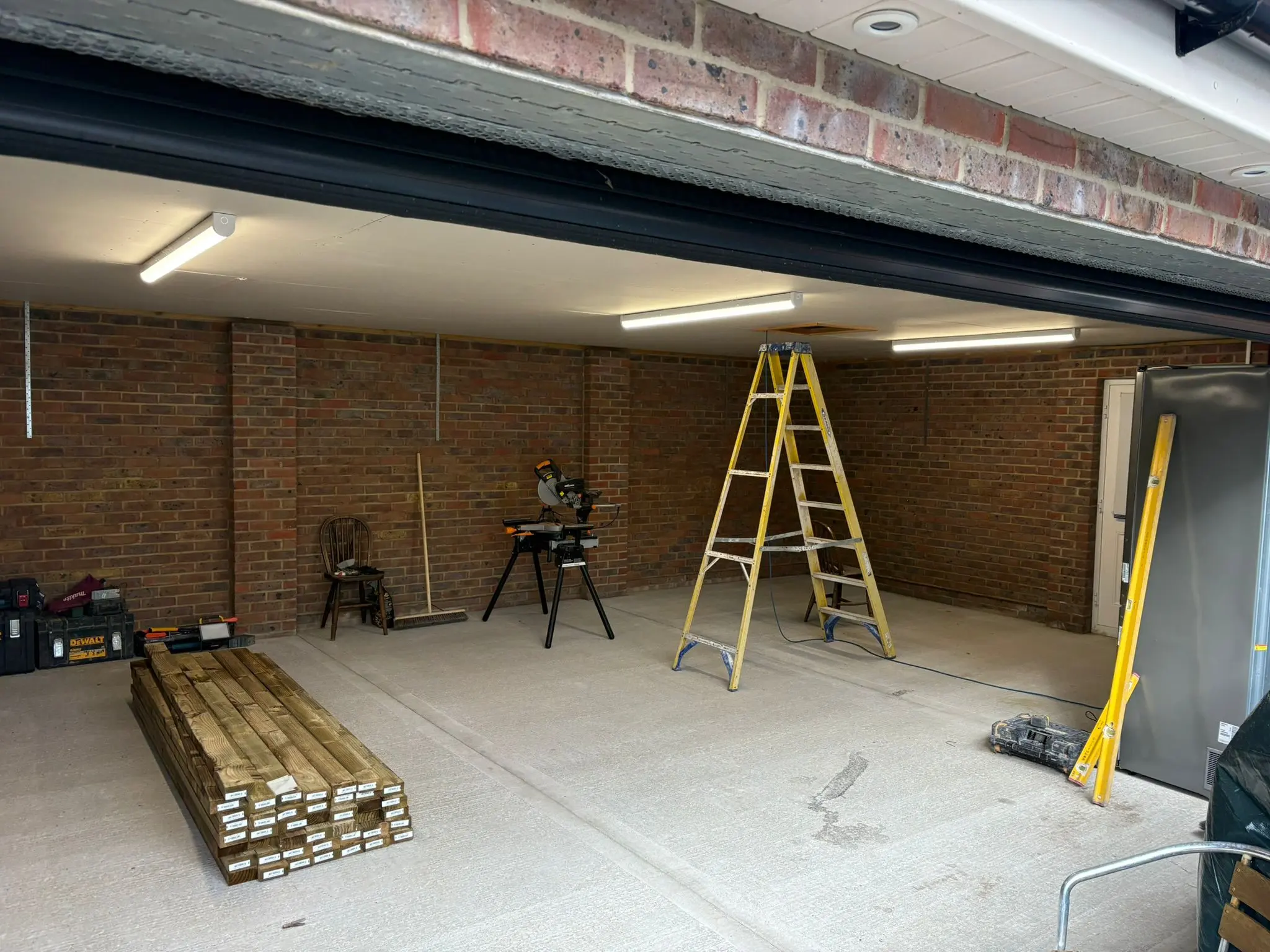 Garage conversion in progress showing exposed brick walls, new ceiling lighting, construction tools, and stacked timber