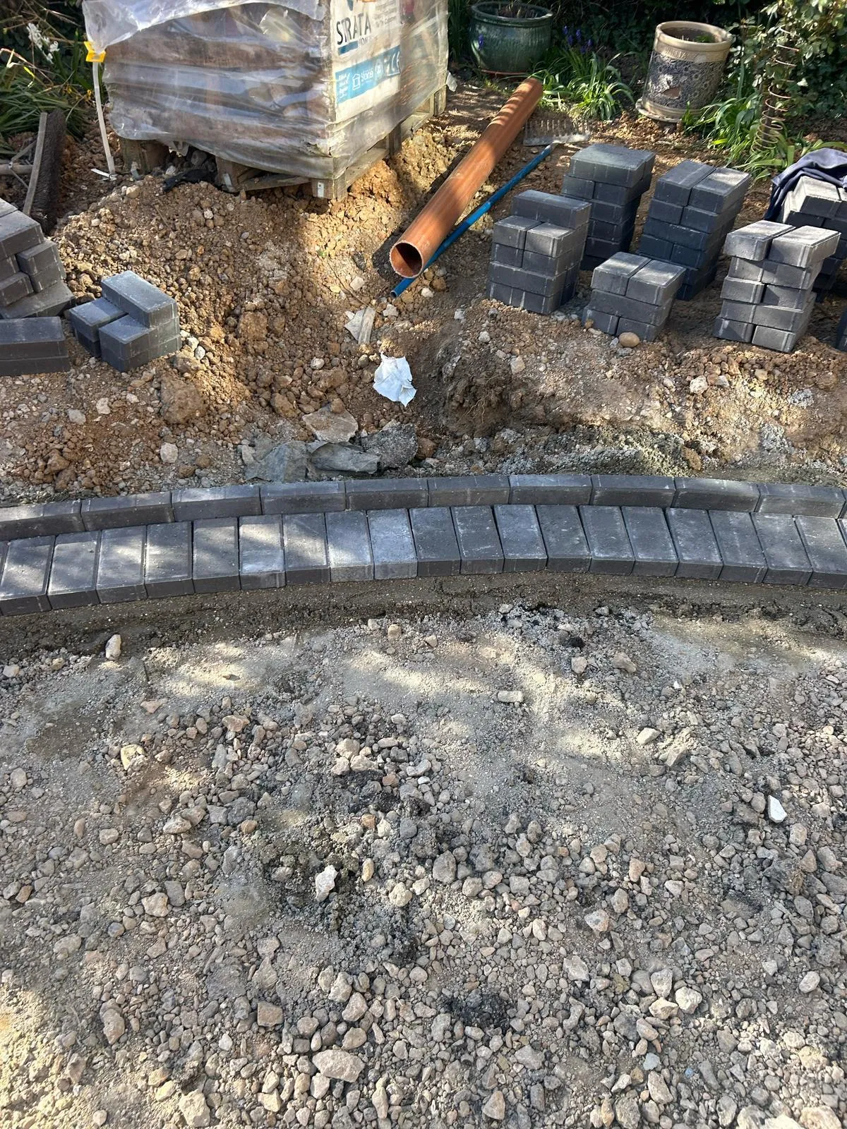 Work in progress on a block paving driveway, showing grey paving blocks forming a curved edge, a gravel sub-base, and construction materials including more blocks and a drainage pipe.