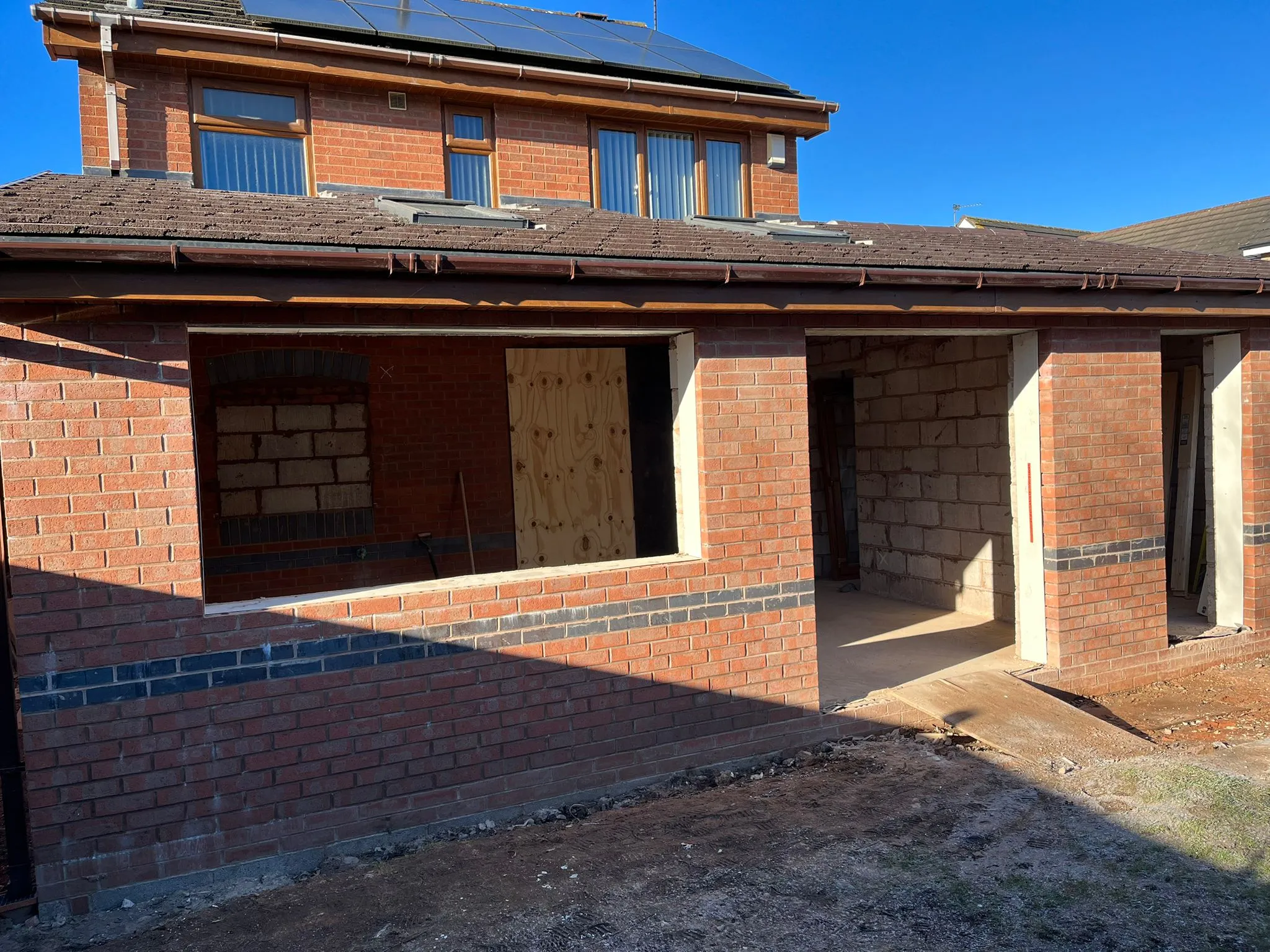 An in-progress home extension with red brick walls, open window and door frames partially boarded up, and a new roofline connecting to the main house.