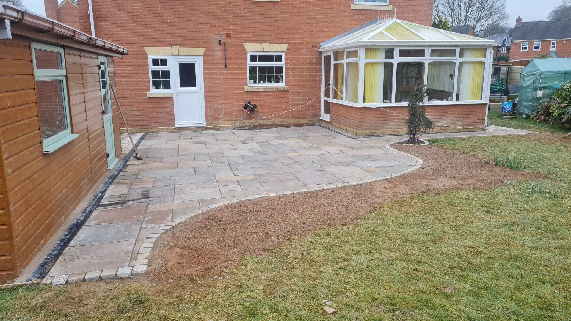 A spacious, newly laid patio made of light-coloured paving slabs with a decorative curved edge of smaller, darker blocks, extending from the back of a brick house with a conservatory to a wooden shed, with a lawn and newly prepared soil in the foreground.