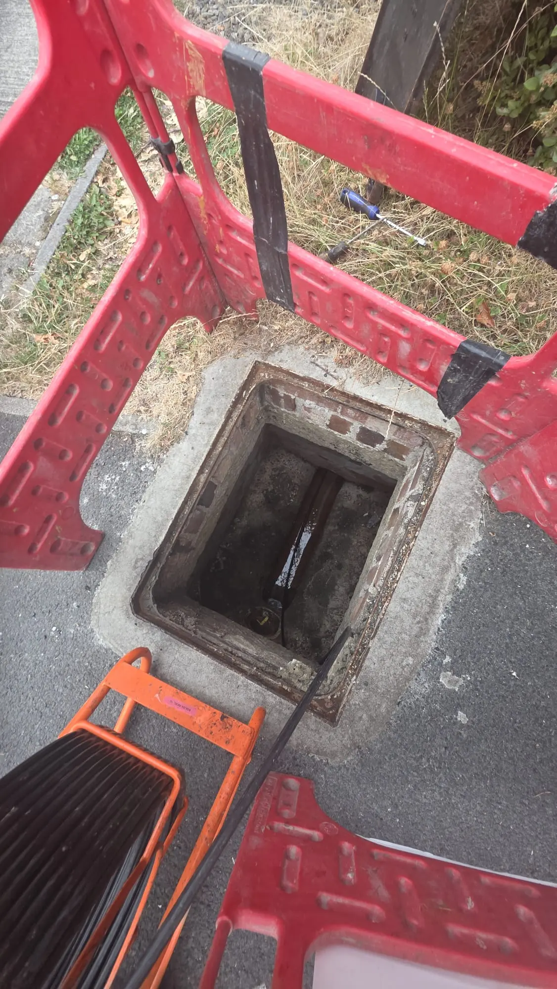 Open manhole surrounded by a red safety barrier with drainage inspection equipment in use.