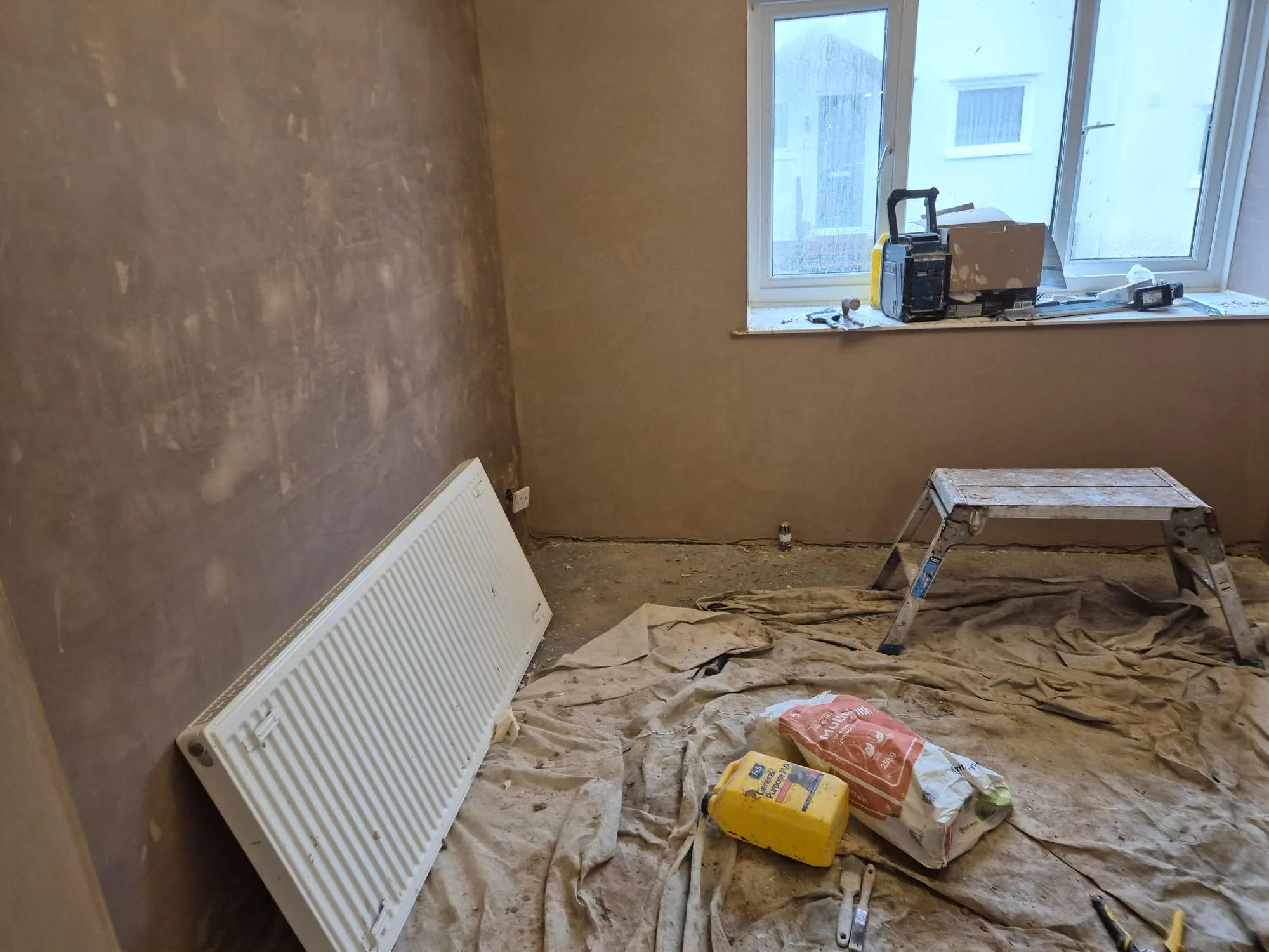 Interior view of a room undergoing renovation, showing newly plastered walls, building materials, tools, and a radiator.