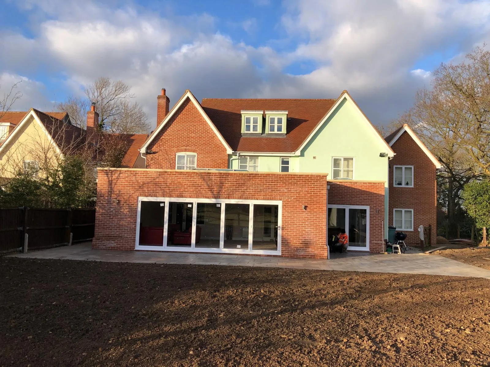 Rear view of a modern house with a new brick extension, white bi-fold doors, and a new grey patio area, viewed from a garden.
