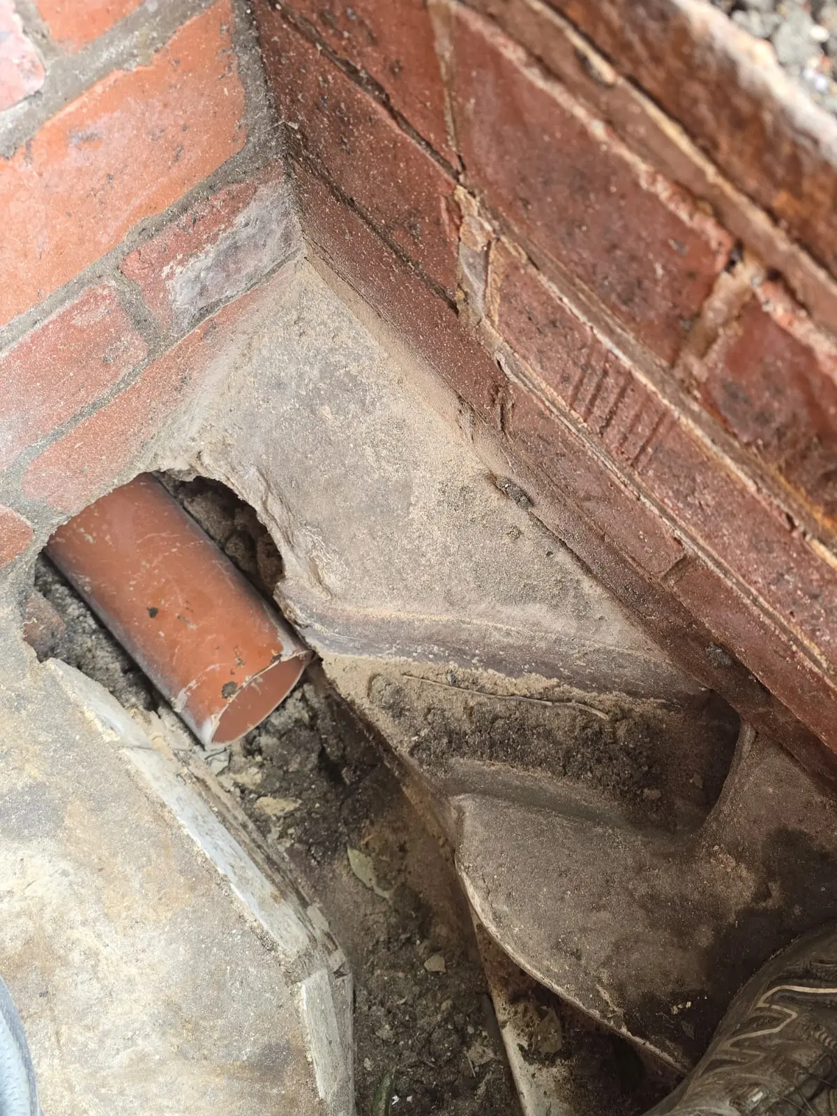 An exposed and broken underground drainage pipe next to a brick wall needing structural repair