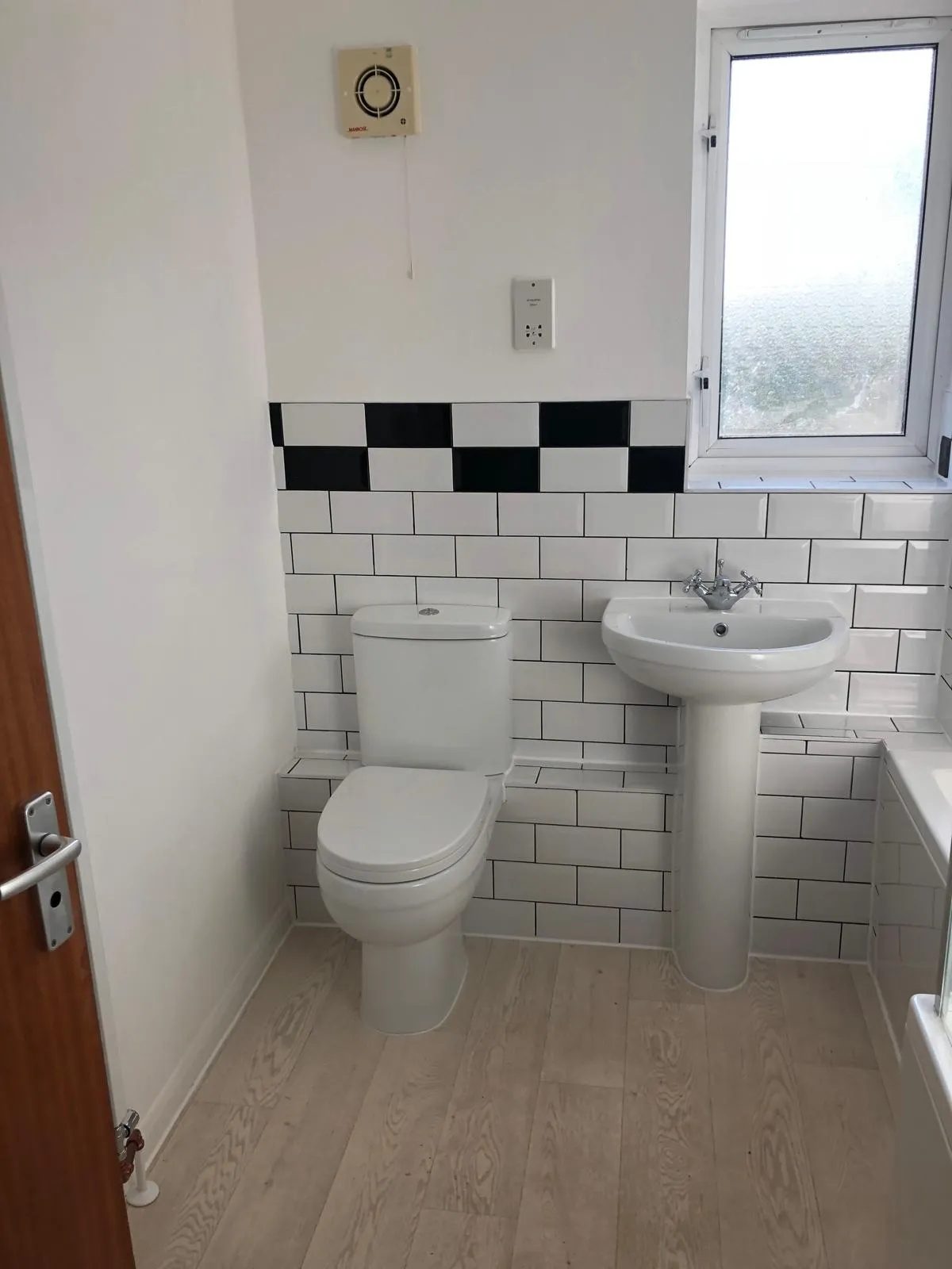 Newly renovated bathroom with white subway tiles, a black and white checkerboard border, toilet, pedestal sink, and light wood-effect flooring.