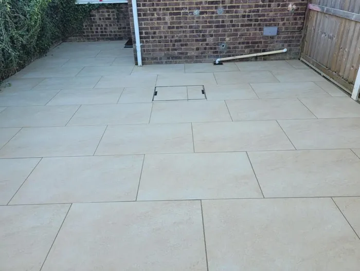 A newly laid modern patio with light-coloured rectangular porcelain-style paving slabs, featuring an integrated drainage cover next to a brick wall and wooden fence.