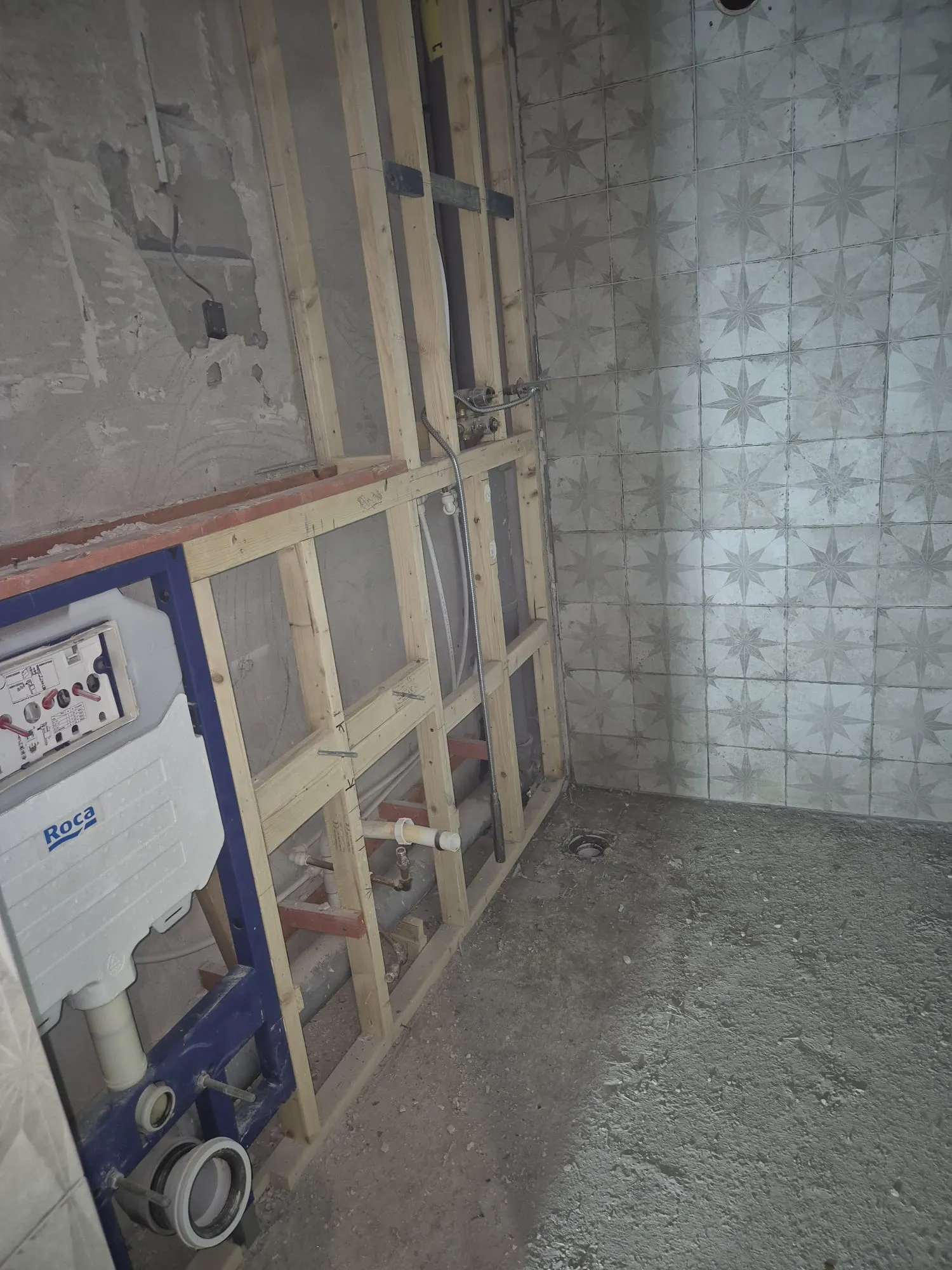 Bathroom renovation in progress with timber stud wall, exposed plumbing, Roca toilet cistern frame, and patterned wall tiles.