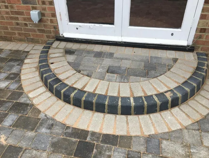 Semicircular block paved steps and patio in light and dark grey, leading up to a white door on a brick house.