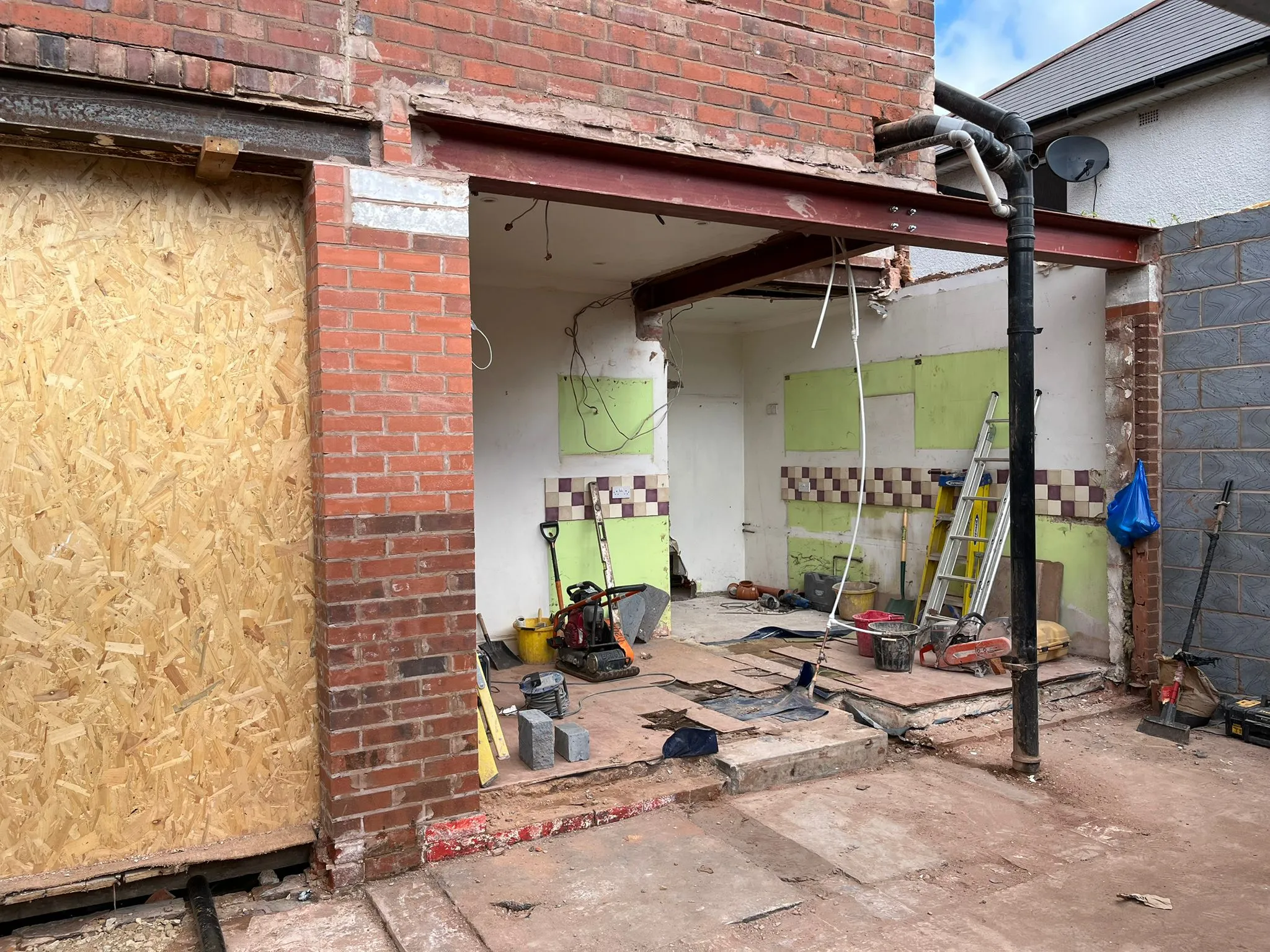 Home extension construction site with large steel beams installed to create an open-plan area, featuring brickwork, boarded-up sections, and building equipment.