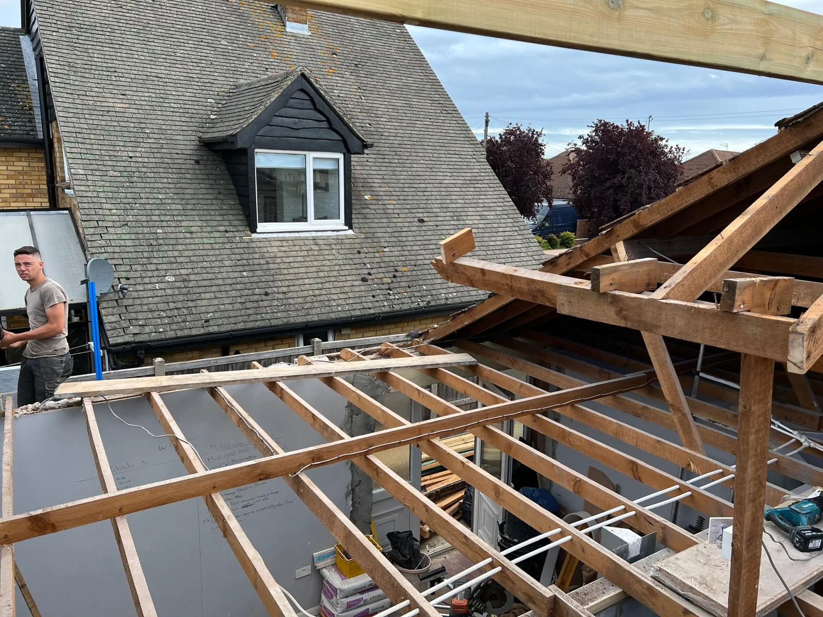 A construction site of a house extension with exposed timber roof joists and a builder, alongside an existing house with a dormer window.