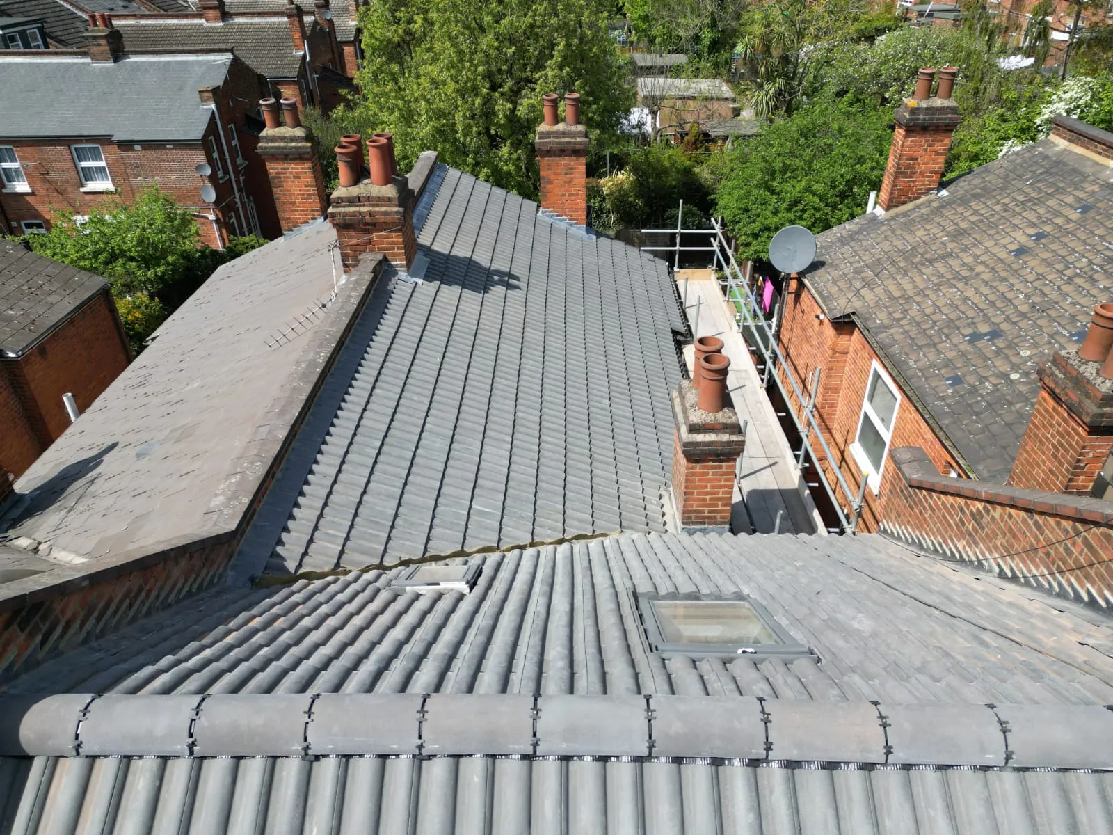 Pitched tiled roofs on residential properties, featuring a newly installed roof section, multiple chimneys, roof windows, and scaffolding, indicating ongoing roofing services.