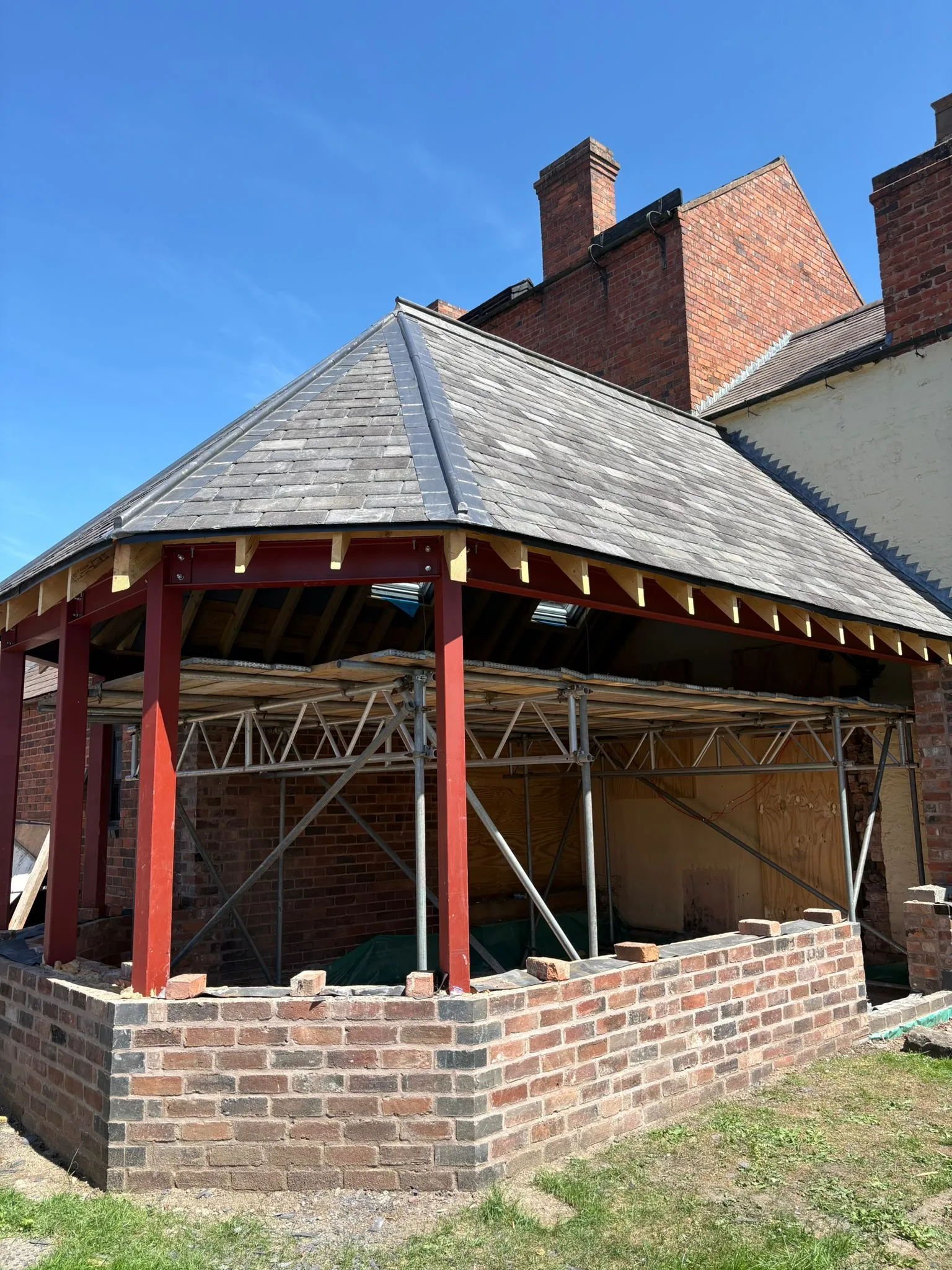 An octagonal home extension under construction, showing the grey tiled roof, red structural steel beams, scaffolding, and new brickwork walls against a clear blue sky.