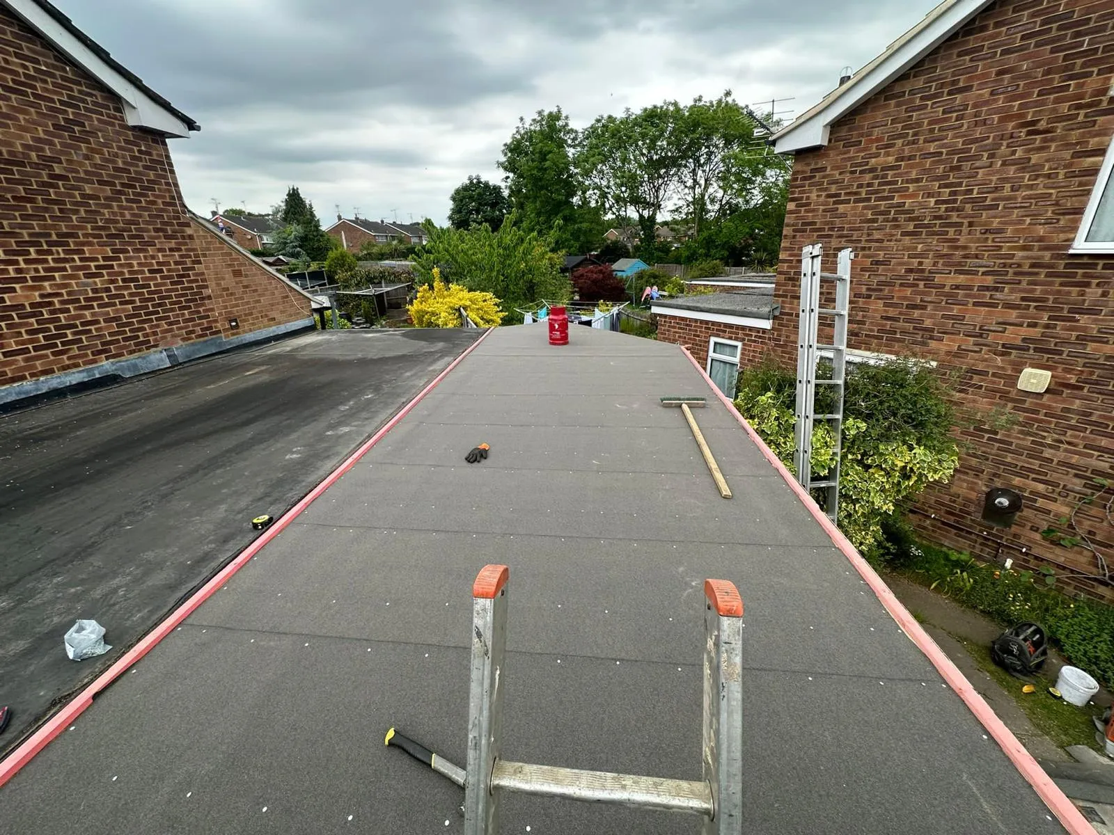 View from a ladder of a newly installed dark grey flat roof, with tools like a broom and canister. An older, black flat roof and two brick houses are visible on either side, with green gardens and an overcast sky in the background.