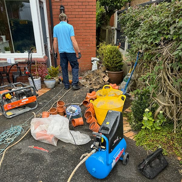 Professional drain excavation and repair work in progress at a residential property showing a technician, exposed trench, and new drainage pipes.
