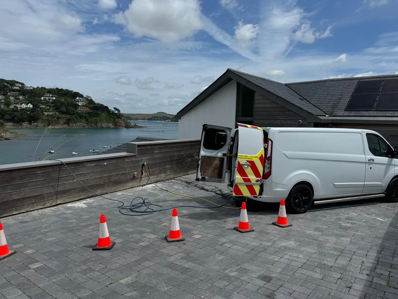 Devon and Cornwall Drainage service van with high-pressure water jetting equipment and hoses, parked at a modern house overlooking a bay, ready for drain unblocking.