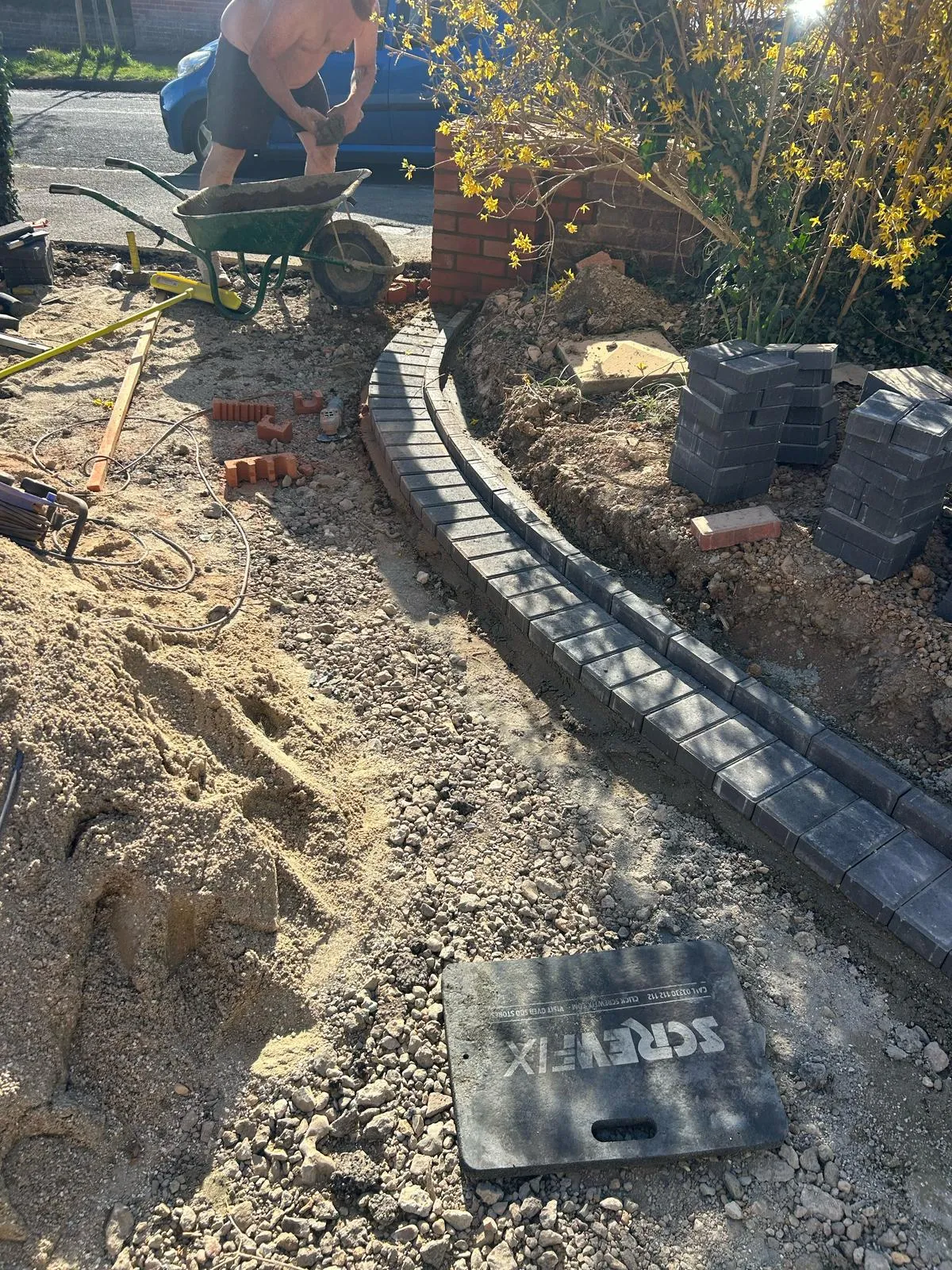 Construction site for a new driveway or garden path, with a worker in the background and a curved border of dark grey block paving being laid. Sand, gravel, and stacks of paving blocks are visible, indicating groundwork and installation.