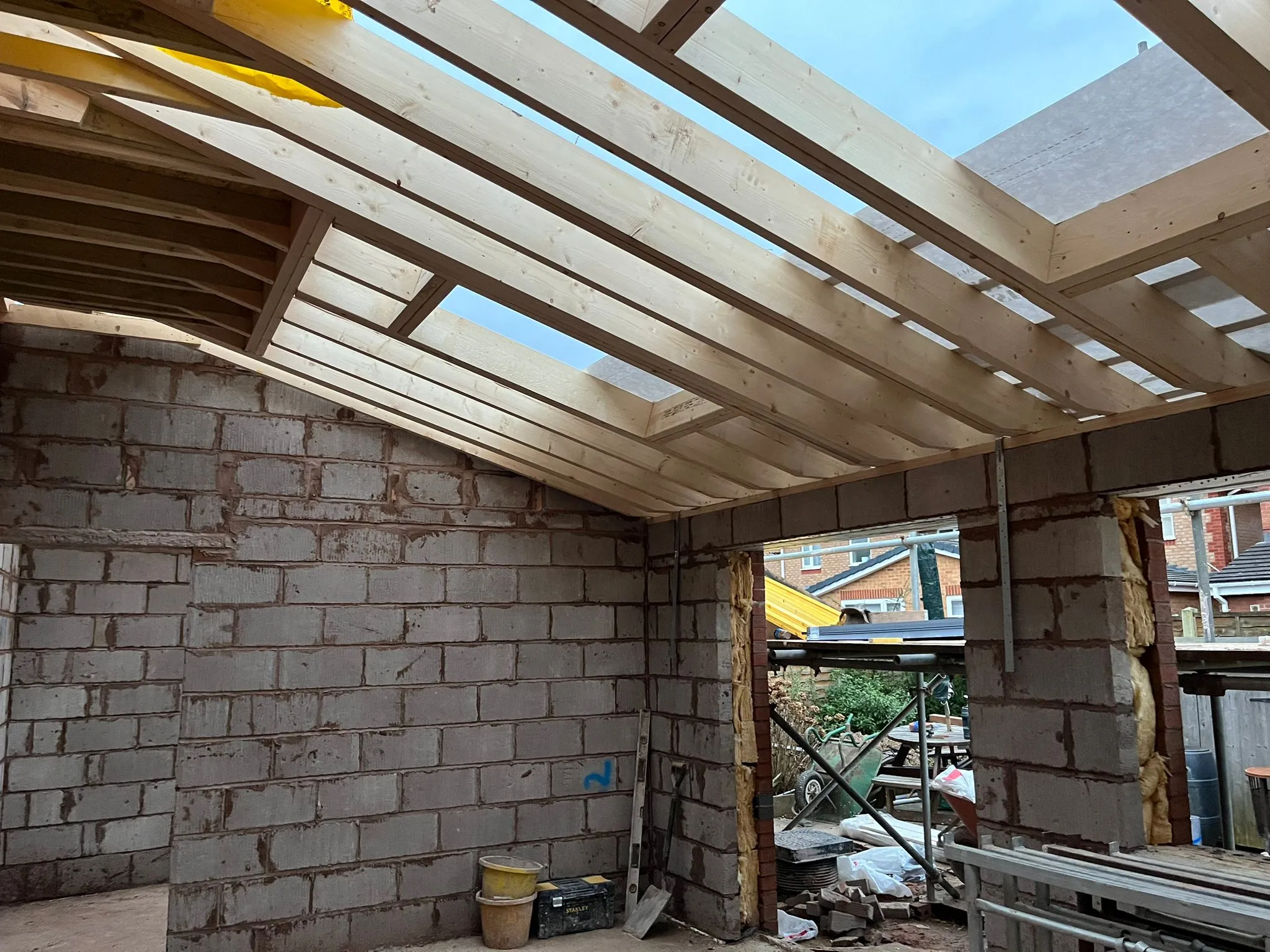 Inside a home extension during construction, showing newly laid grey block walls, exposed wooden roof joists, and open gaps for skylights against a cloudy sky.