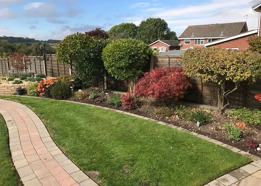 A professionally maintained domestic garden with a curved block paving path, green lawn, and landscaped planted beds, under a bright sky.