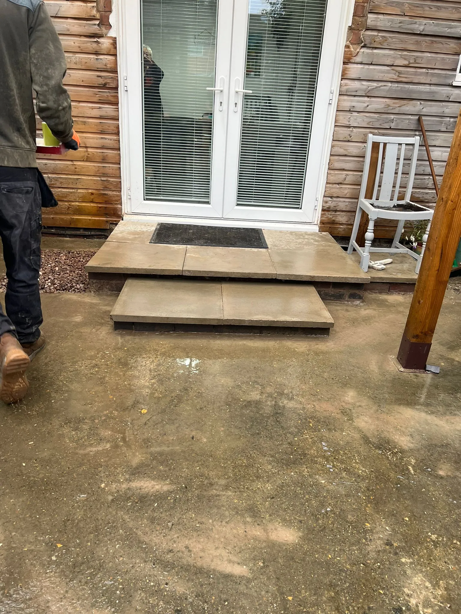 Newly installed paved patio steps against a timber-clad house with white double doors, and a wet ground area in front, indicating recent landscaping work.