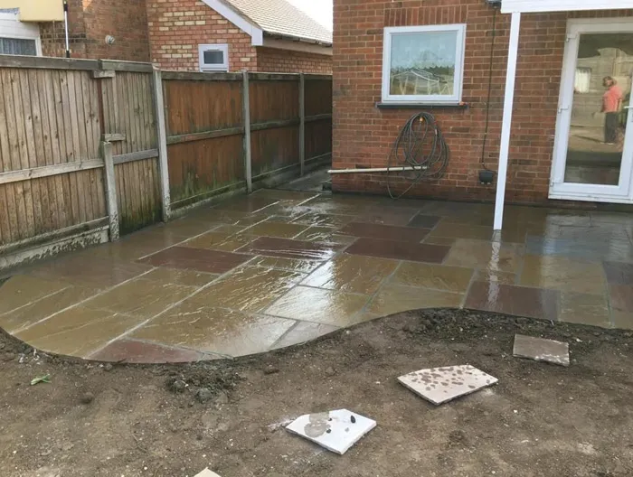 Wet Indian Sandstone patio with mixed colours and a curved border, professionally laid beside a house and a wooden garden fence, highlighting new paving work.