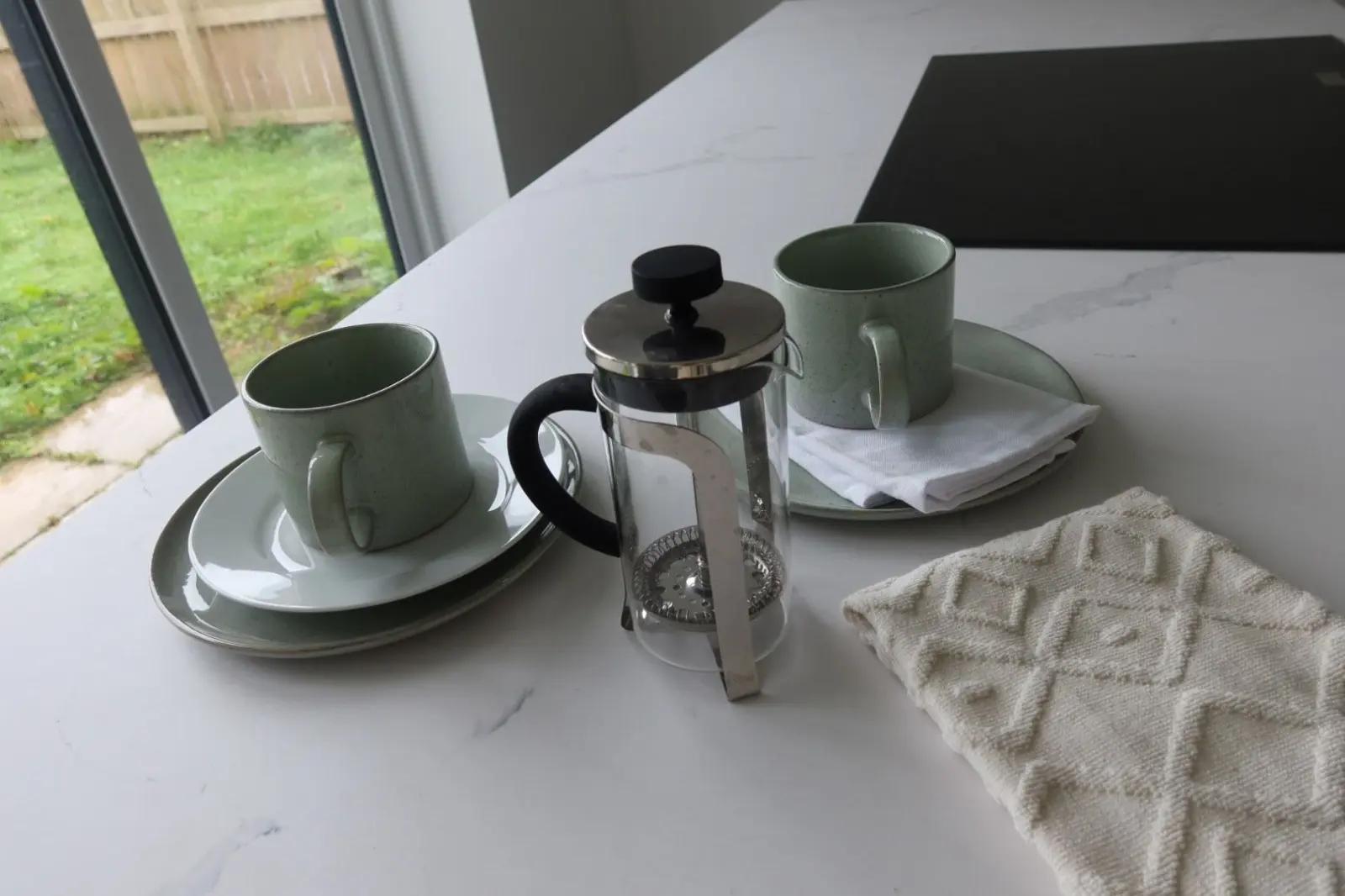 A French press and two coffee mugs on a bright kitchen counter in a warm, comfortable home
