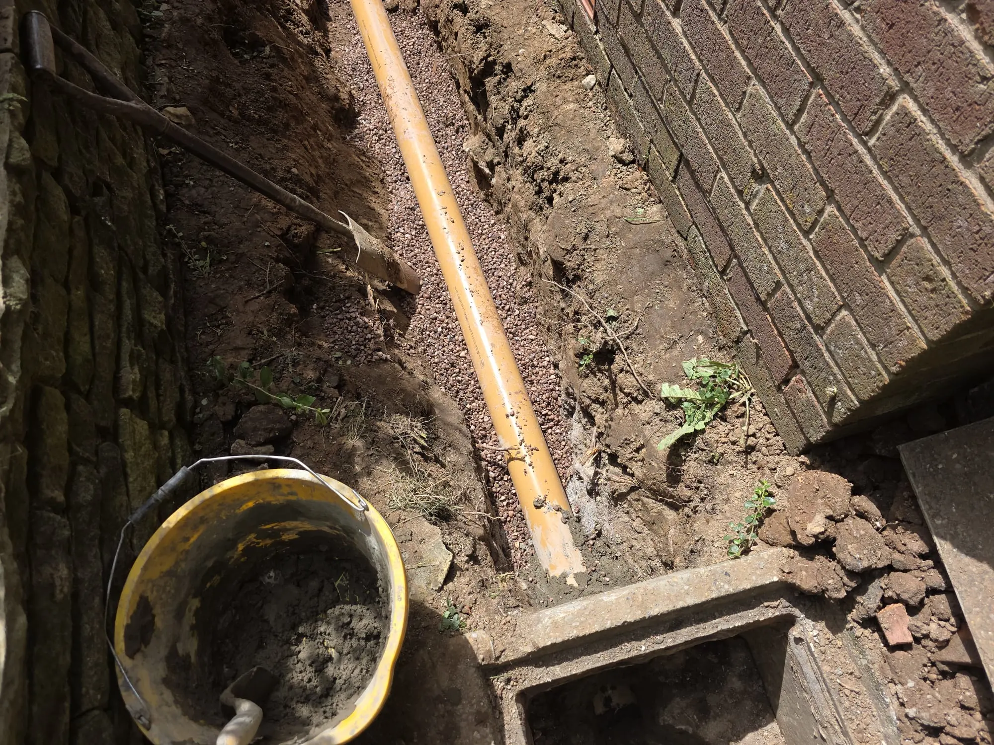 Newly laid orange drainage pipe in an excavated trench with gravel bedding alongside a brick wall