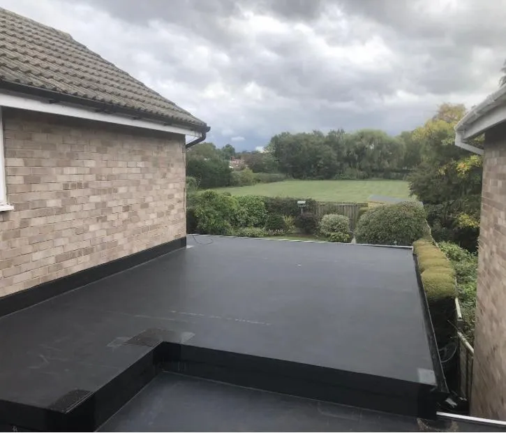 A completed flat roof installation on a home extension, showing the sleek, waterproof surface alongside the main tiled roof of the house and a distant field.