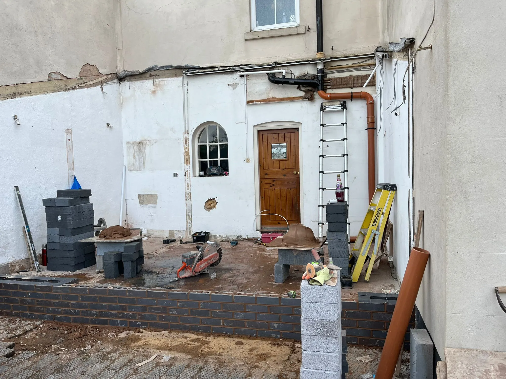 Construction site showing the start of a home extension with new brick foundations, building materials, and tools, next to an existing house wall.
