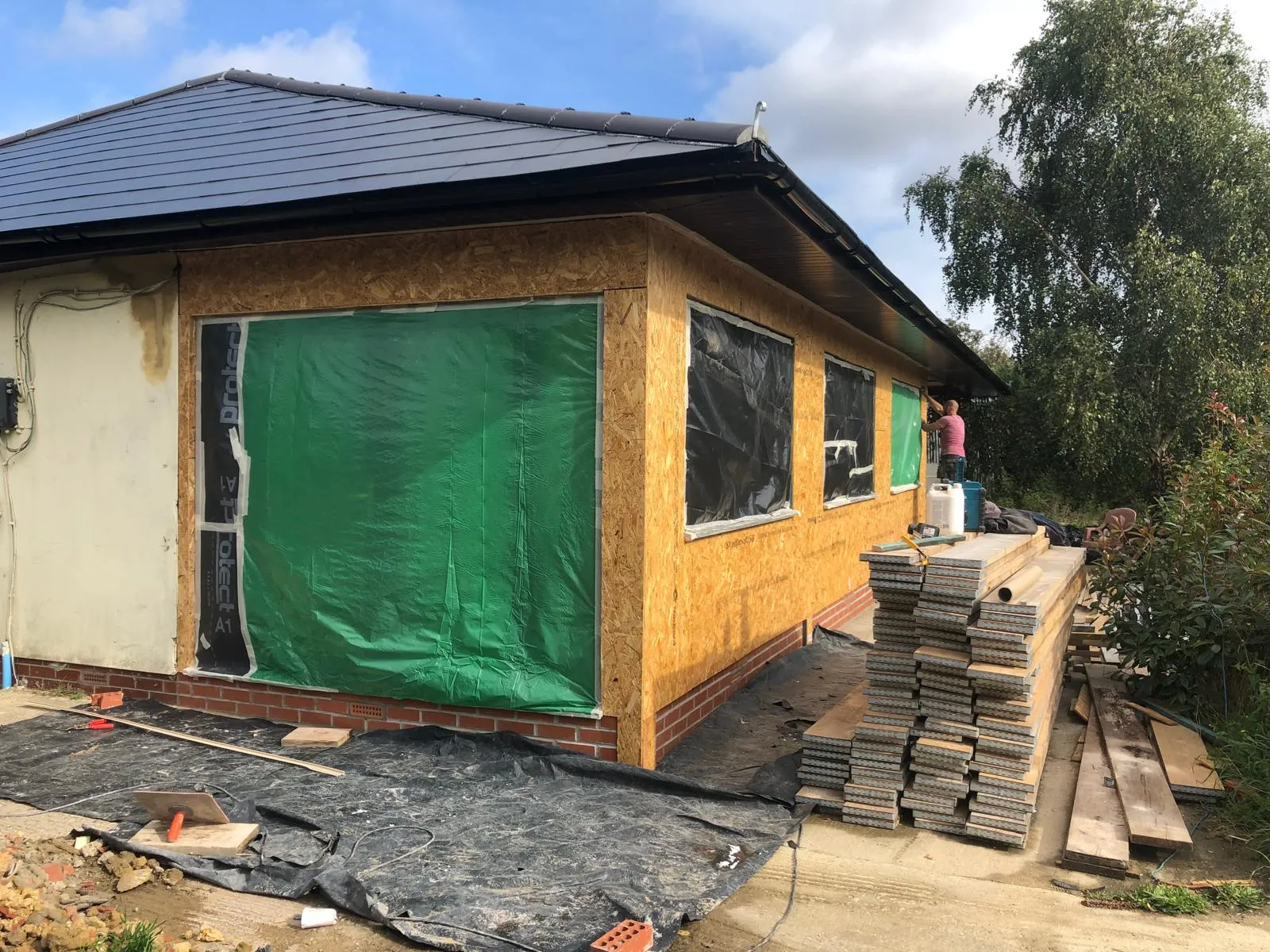 A building under construction, likely a house extension, with OSB sheathing on walls and a dark tiled roof, with construction materials and a worker visible.