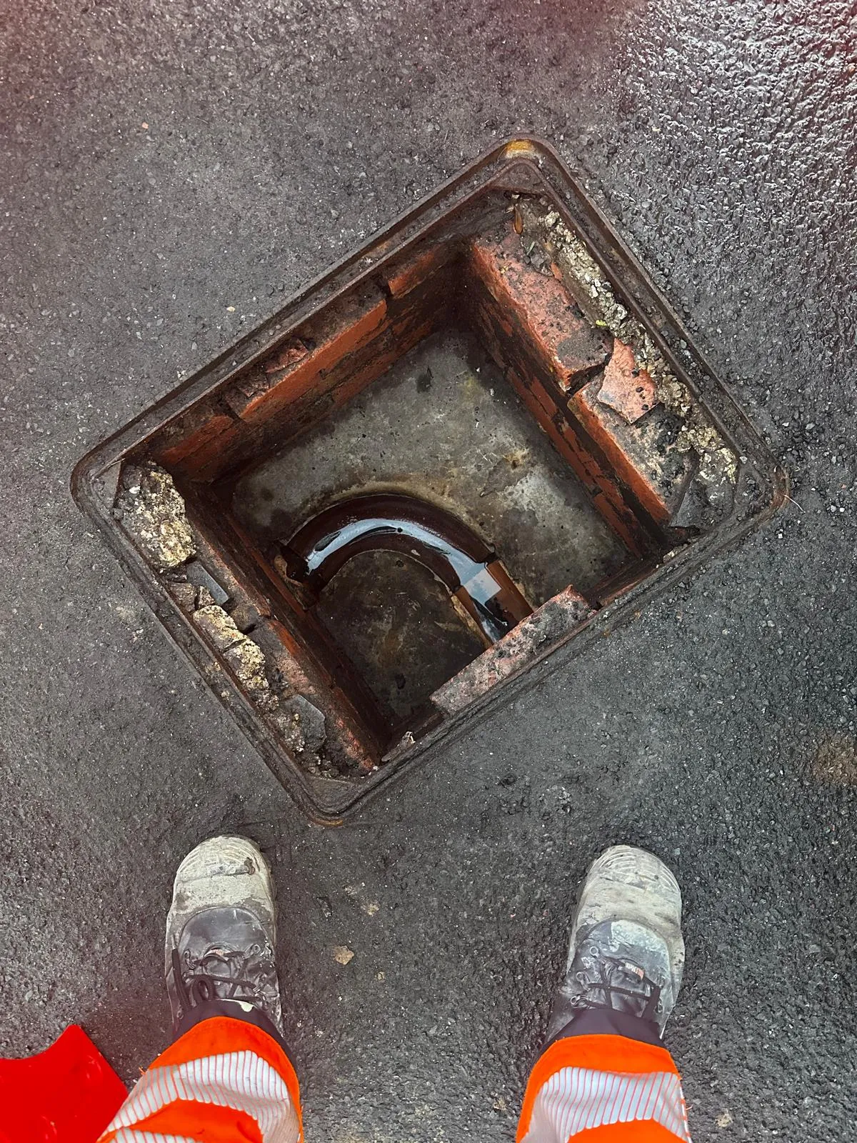 A close-up of a drainage professional's feet and an open manhole chamber with a u-bend pipe containing water, on an asphalt surface.