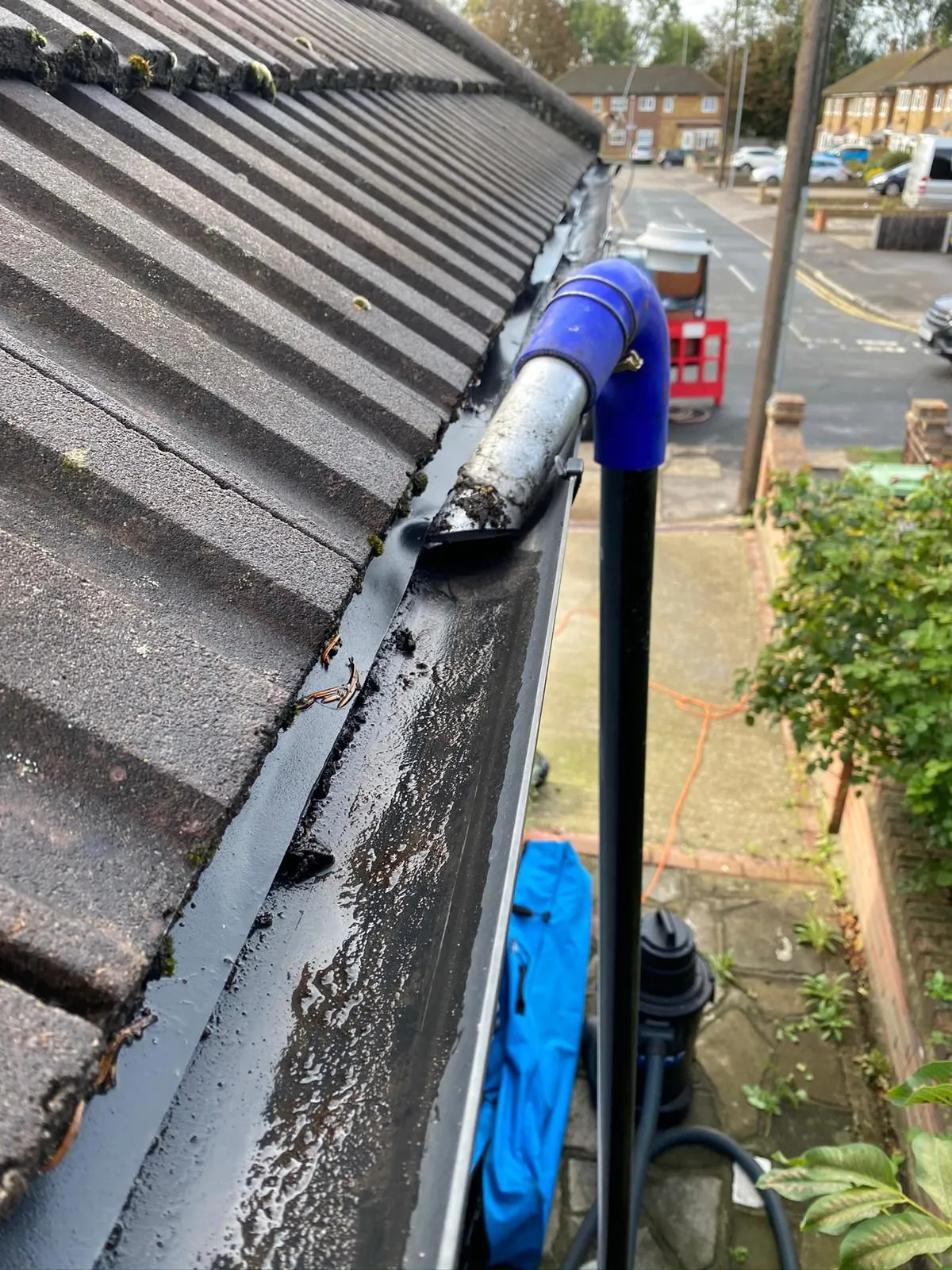 A high-reach vacuum tool being used to clean out a black gutter on a tiled roof.