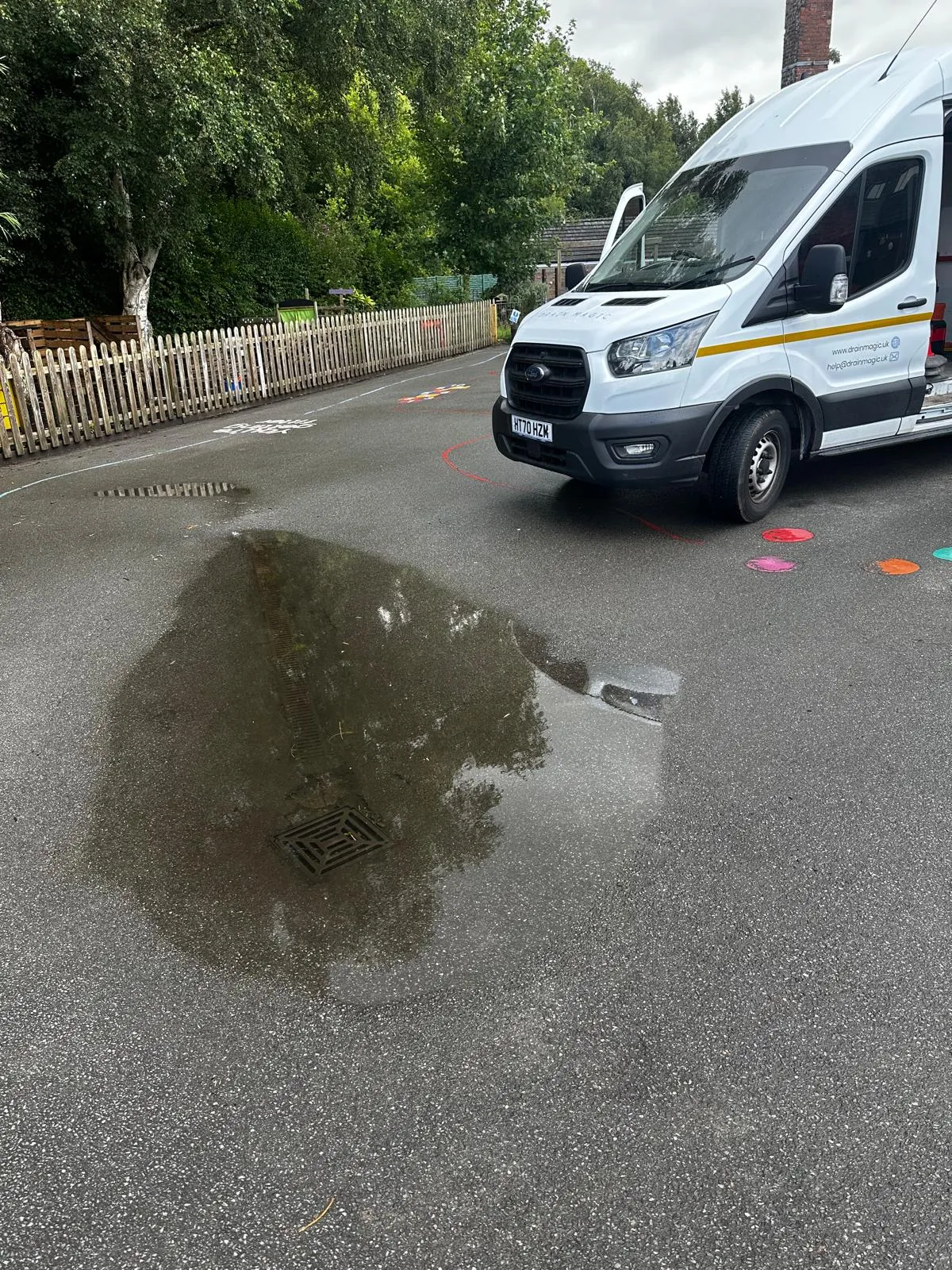 An outdoor blocked drain causing a large puddle on tarmac, with a white service van ready to clear the blockage, suggesting an emergency call-out.