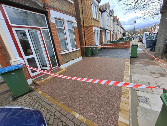 A close-up view of a newly installed resin-bound driveway, featuring light brown aggregate, in front of a brick terraced house. Red and white barrier tape is stretched across the drive to indicate it is new. A green refuse bin is visible to the left.