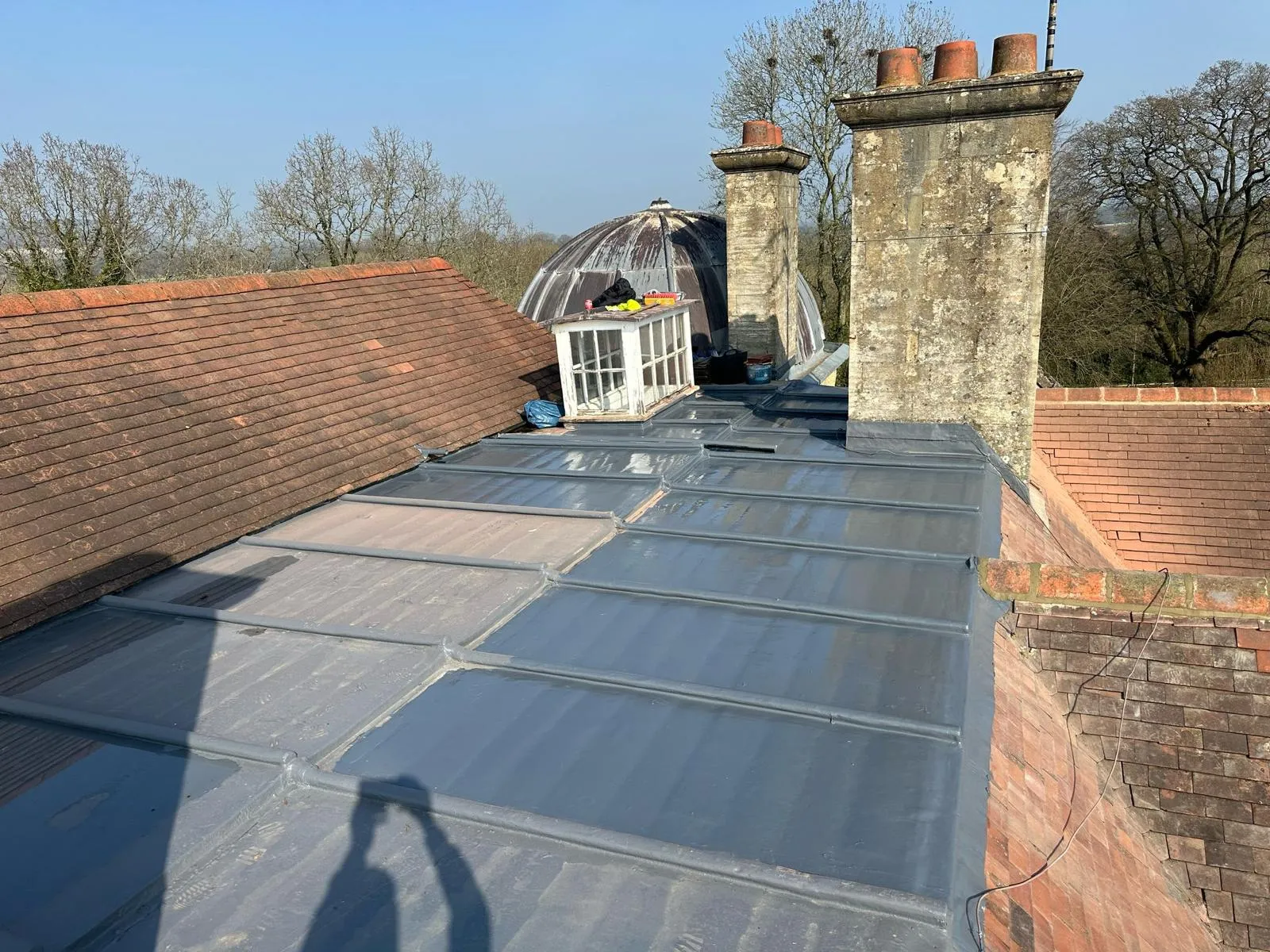 Residential roof with new grey metal standing seam, old terracotta tiles, brick chimneys, and a small roof light.