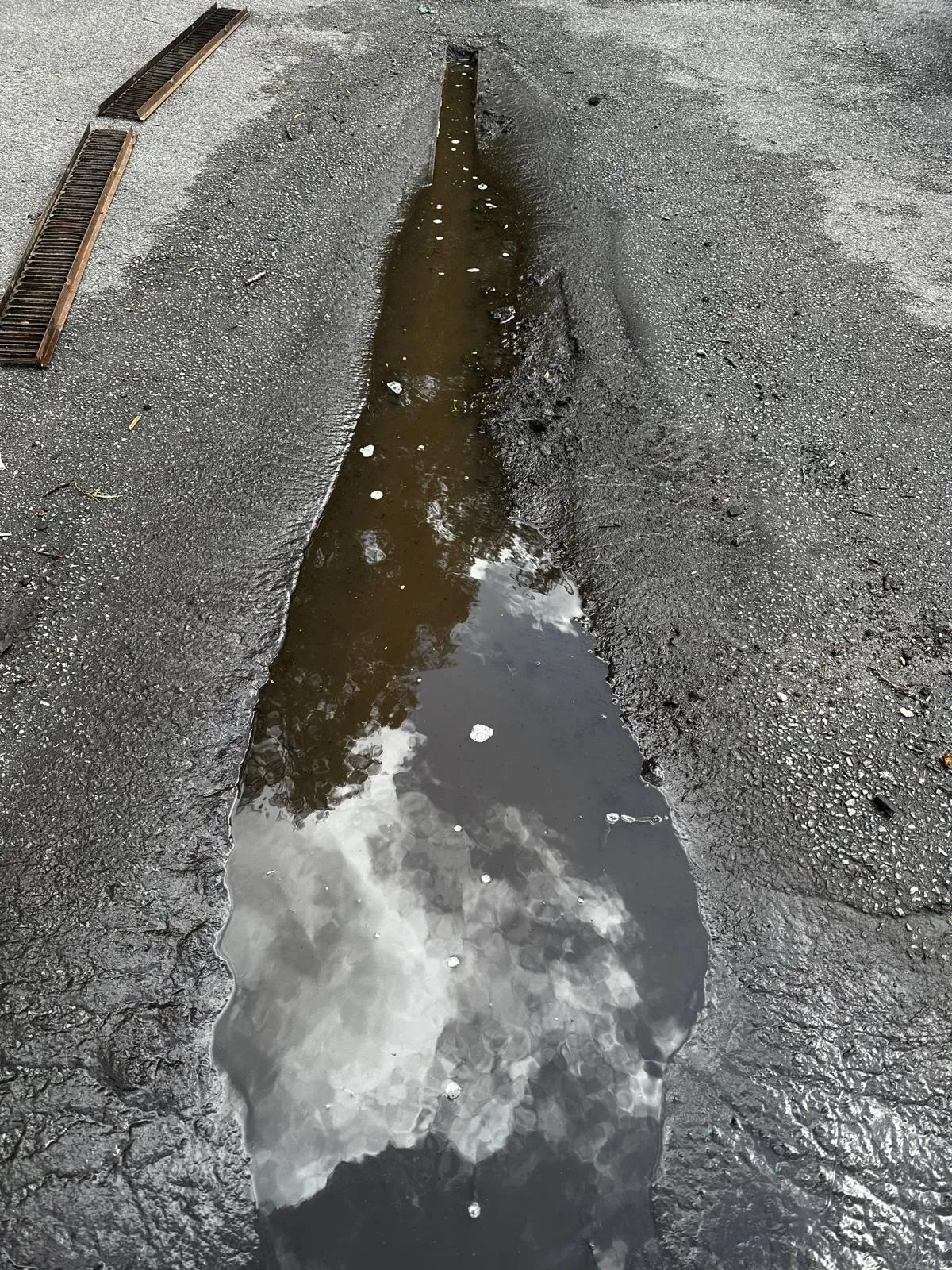 Overflowing external drain or gully with dirty water and two displaced metal grates, reflecting clouds.