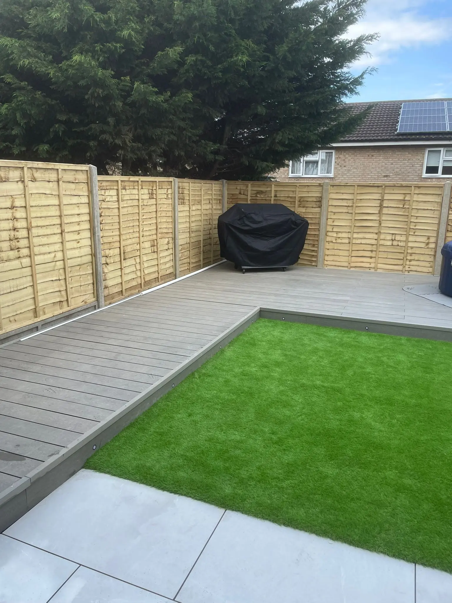 Modern garden landscaping featuring grey composite decking, artificial grass, porcelain patio slabs, and new fencing installed in a residential backyard.