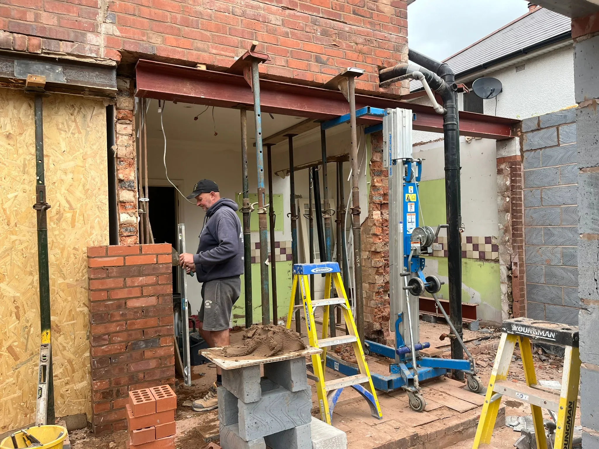 A builder laying bricks on a new home extension construction site, with steel beams and acrow props supporting the structure.
