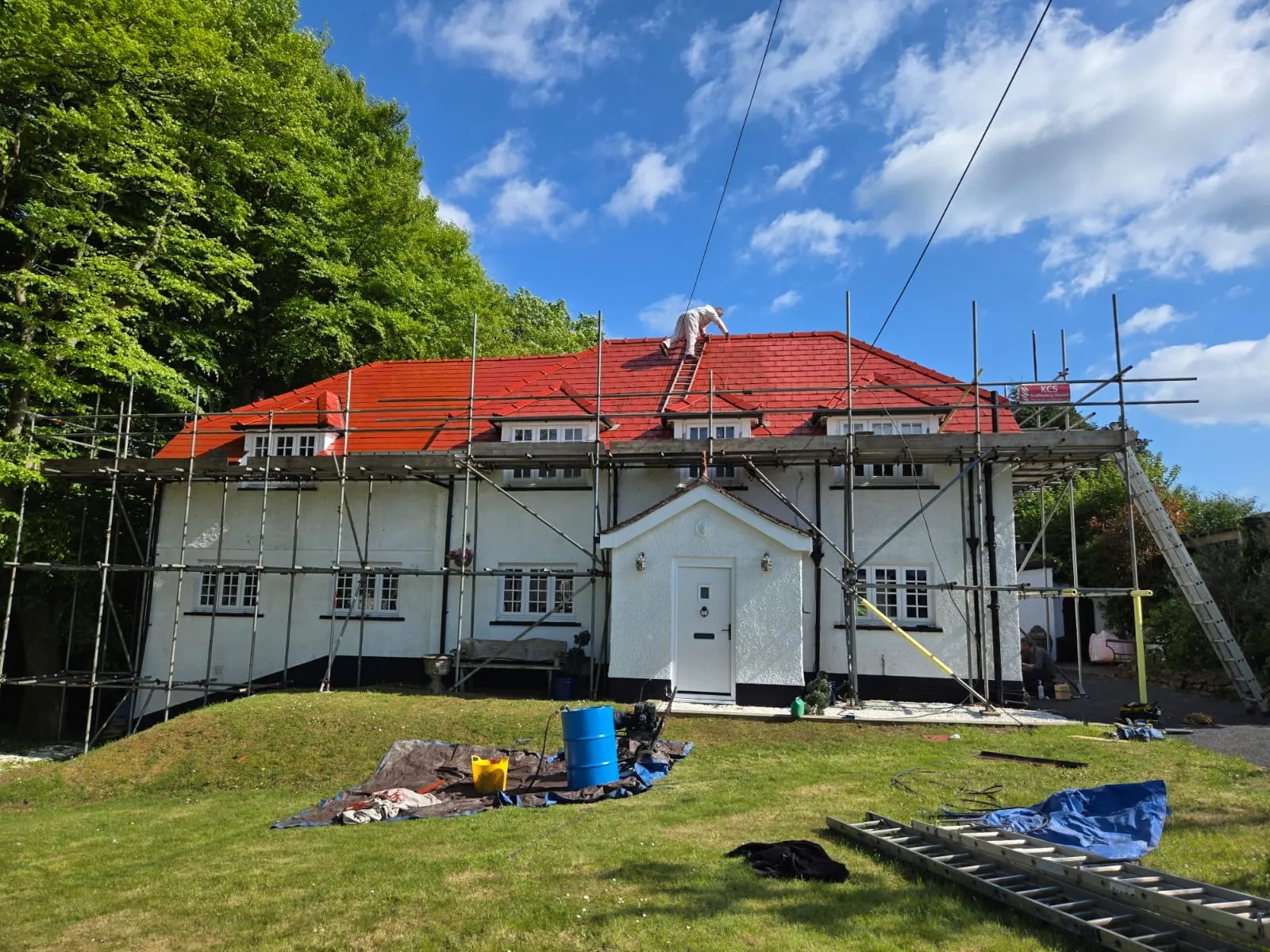 A large white house with a vibrant red roof under renovation, surrounded by scaffolding with a worker on the roof.