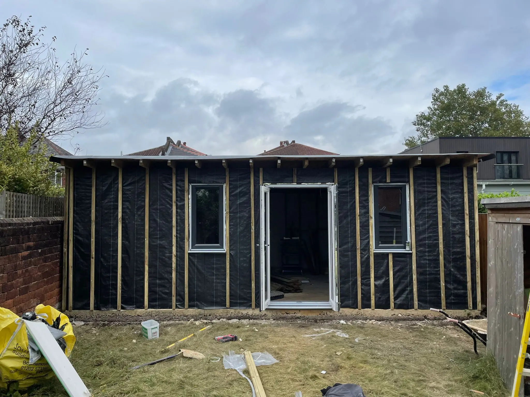 A bespoke timber-framed garden room under construction, showing breathable membrane, vertical battens, and installed windows and French doors in a residential garden.