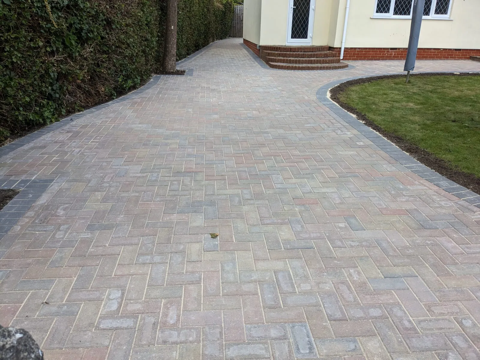 Newly laid block paving driveway with a herringbone pattern, multi-coloured blocks, and a dark kerb, curving around a front garden.