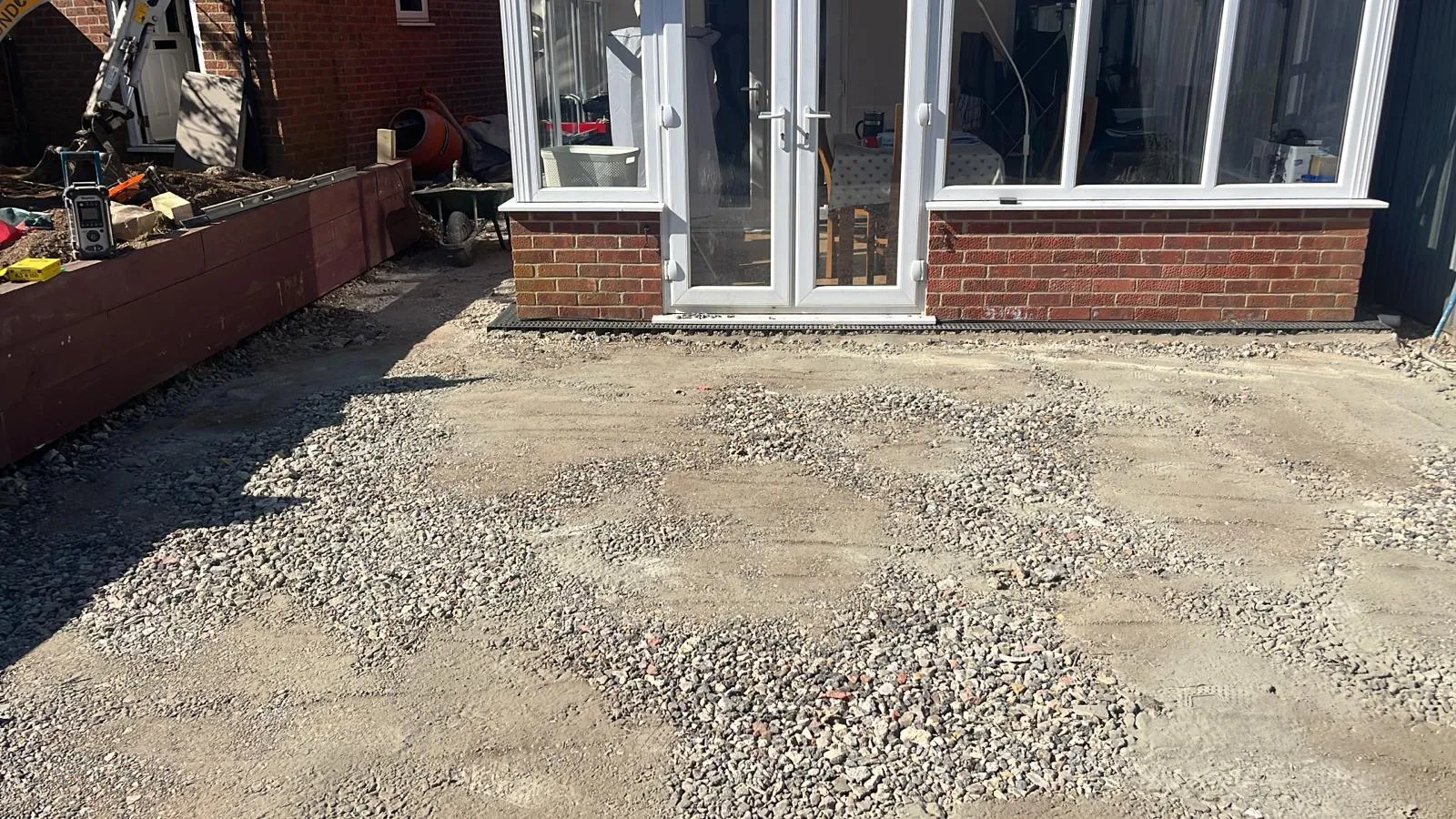 Image depicting ground preparation for a new patio or driveway, with a drainage system in place and aggregate sub-base material spread across the site, ready for further surfacing.
