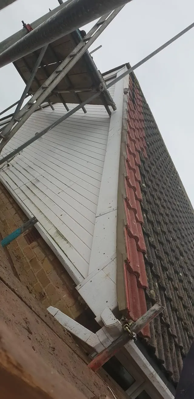 Roof renovation in progress with scaffolding, featuring new red roof tiles being installed alongside old tiles, and white fascias on a gable.