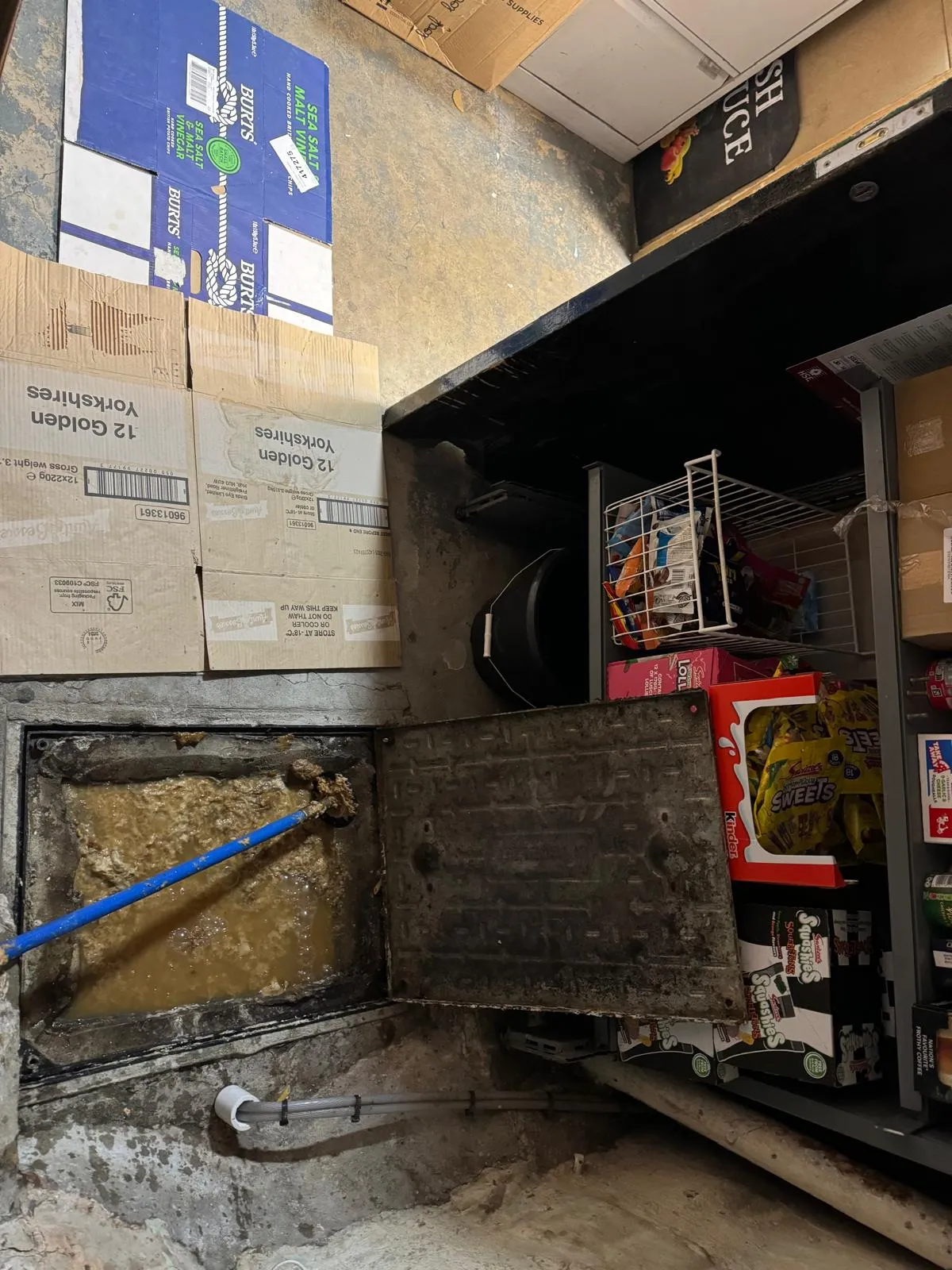 An open drain access point revealing a severe blockage of sludge and dirty water, with a blue drain rod inserted for clearing, surrounded by commercial product boxes.