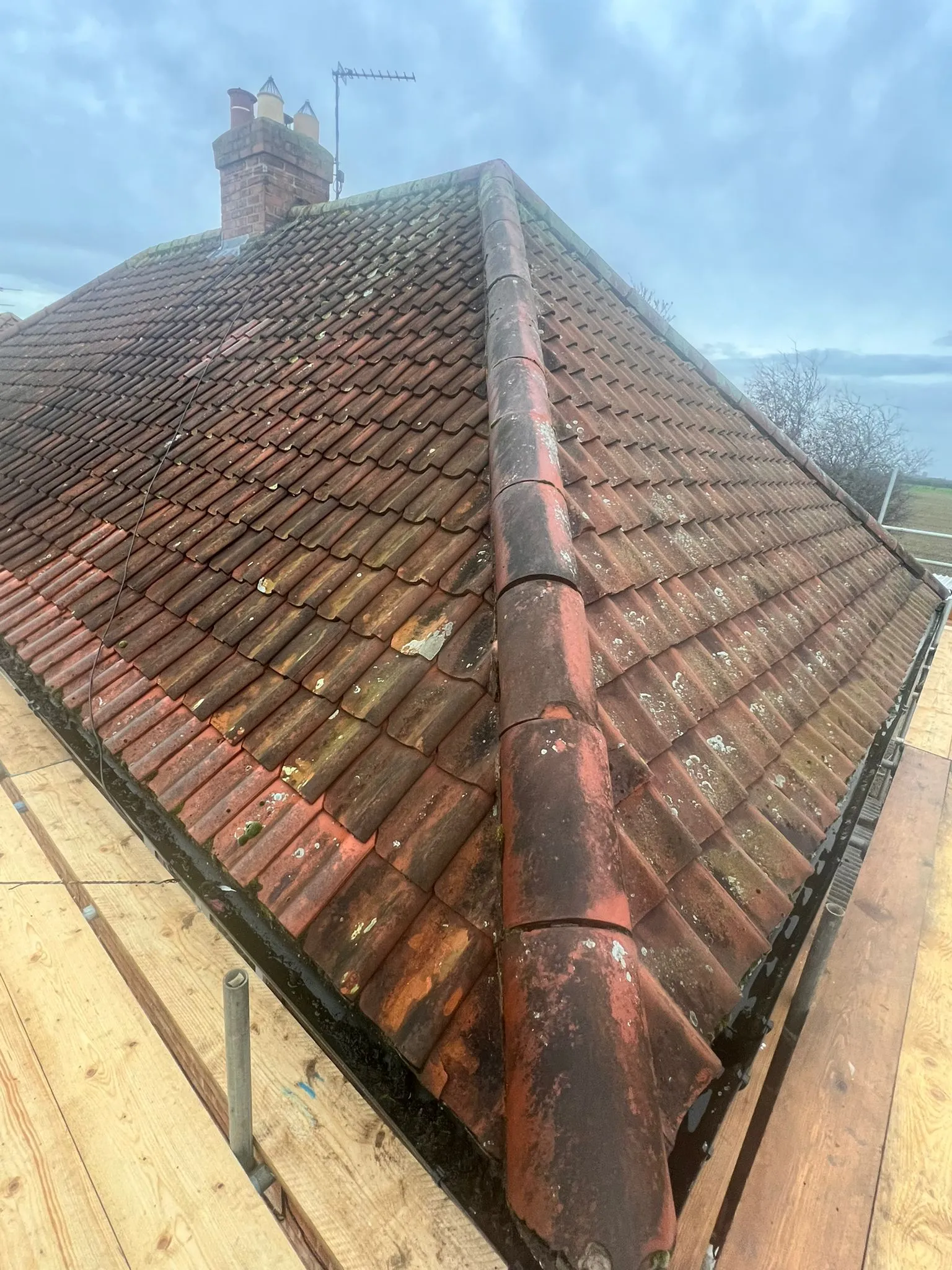 An aged, moss-covered tiled roof with a visible chimney and antenna, seen from scaffolding, highlighting a need for cleaning or repair.
