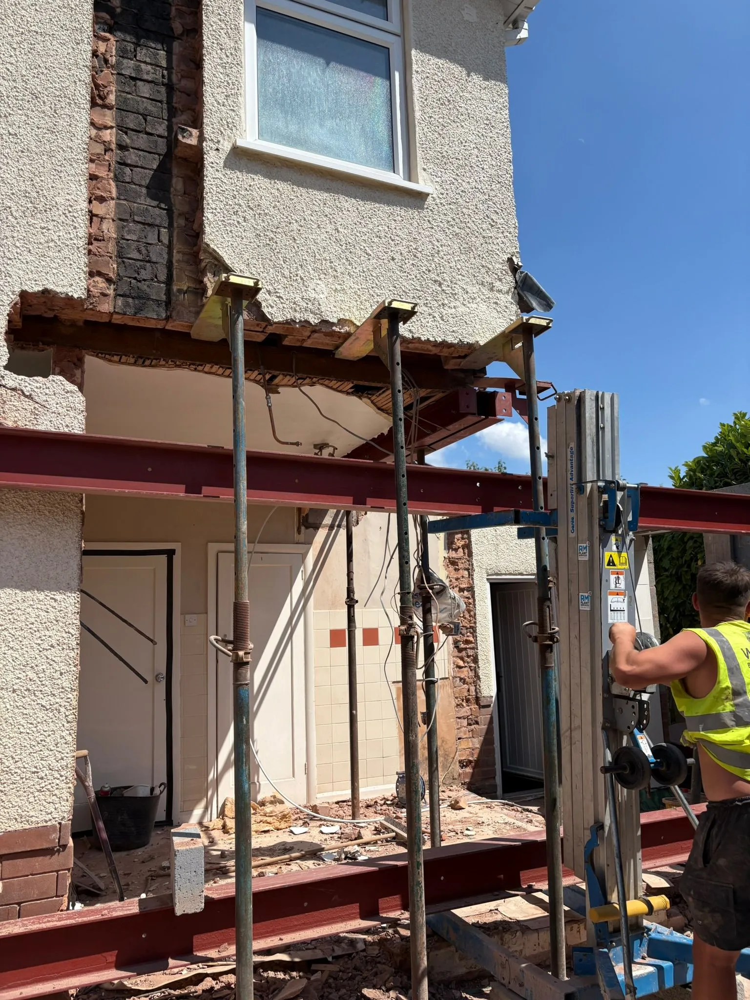 Construction in progress for a home extension, showing steel support beams, scaffolding props, and a worker. The side of a house has a section removed for a new extension.