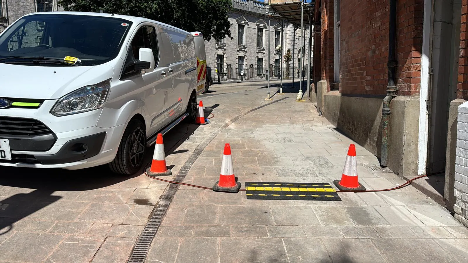 A white drainage service van parked on a street with orange cones and a hose running towards a building, showing an ongoing drain unblocking job.